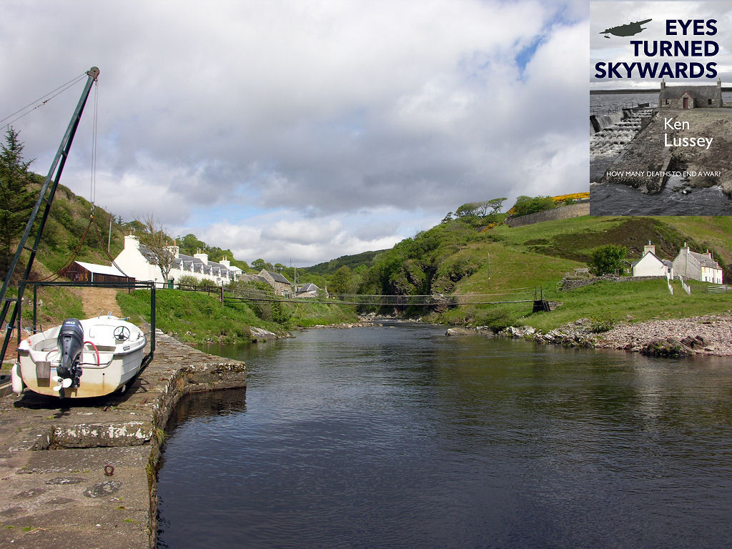 This image shows a view along a river. On the near, left, bank is a quay with a small motorboat under a crane and a row of white cottages beyond it on the side of a slope that climbs a hillside. A pedestrian suspension bridge crosses to the far bank of the river on the right of the frame where there are more cottages with a hillside above them and a stony beach beneath them. The river narrows as it reaches the centre of the frame in a wooded valley. The front cover of ‘Eyes Turned Skywards’ is shown in the top right corner.