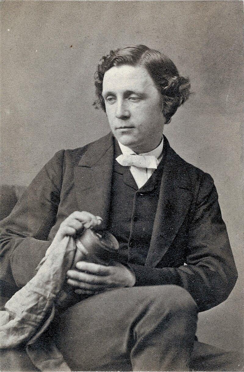 1863 photograph of Lewis Carroll by Oscar G. Rejlander.

The image is a black-and-white portrait showing Carroll, the pen name of Charles Lutwidge Dodgson, dressed formally in a dark suit with a white bow tie. 

He has wavy, neatly styled hair and a calm, contemplative expression. His gaze is directed slightly downward and to the side. 

He is holding an object, possibly a jar or small container, wrapped in a cloth, adding a slightly mysterious element to the composition.