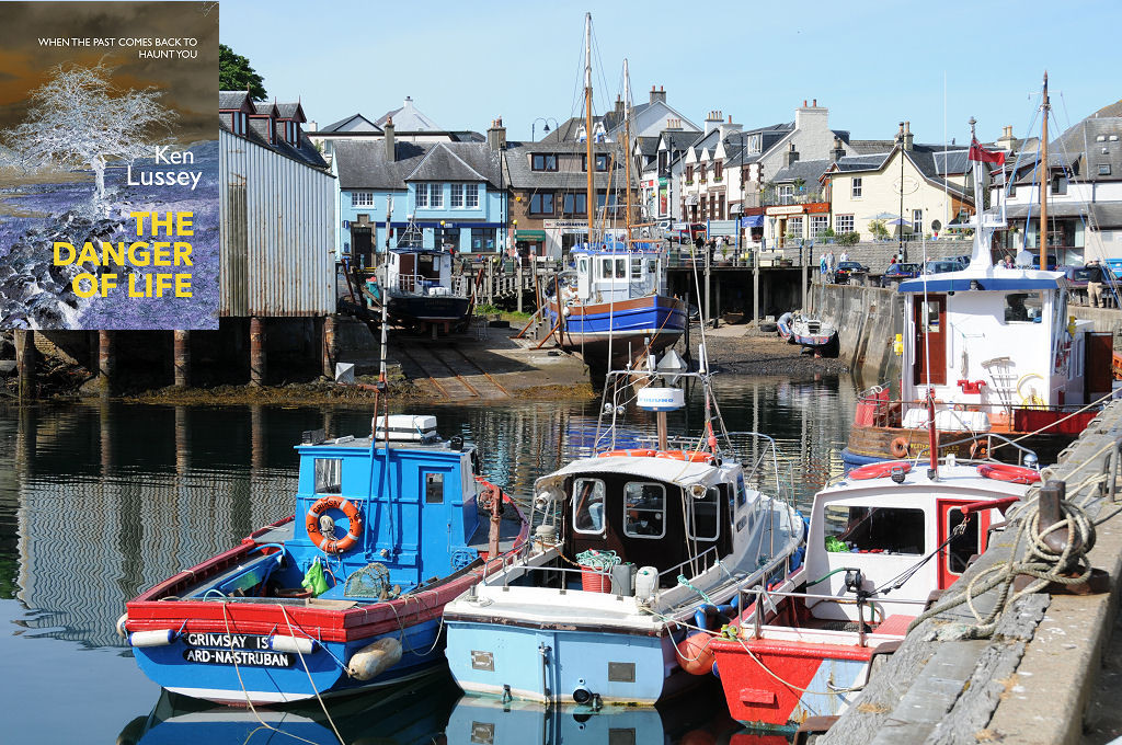 The image shows a view from a pier across a busy harbour towards the village of Mallaig. Small fishing boats are moored up to three abreast and there is a boatyard with slipways beyond them, with boats being repaired and a large shed in the upper left of the frame. The front cover of ‘The Danger of Life’ is shown in the top left corner.