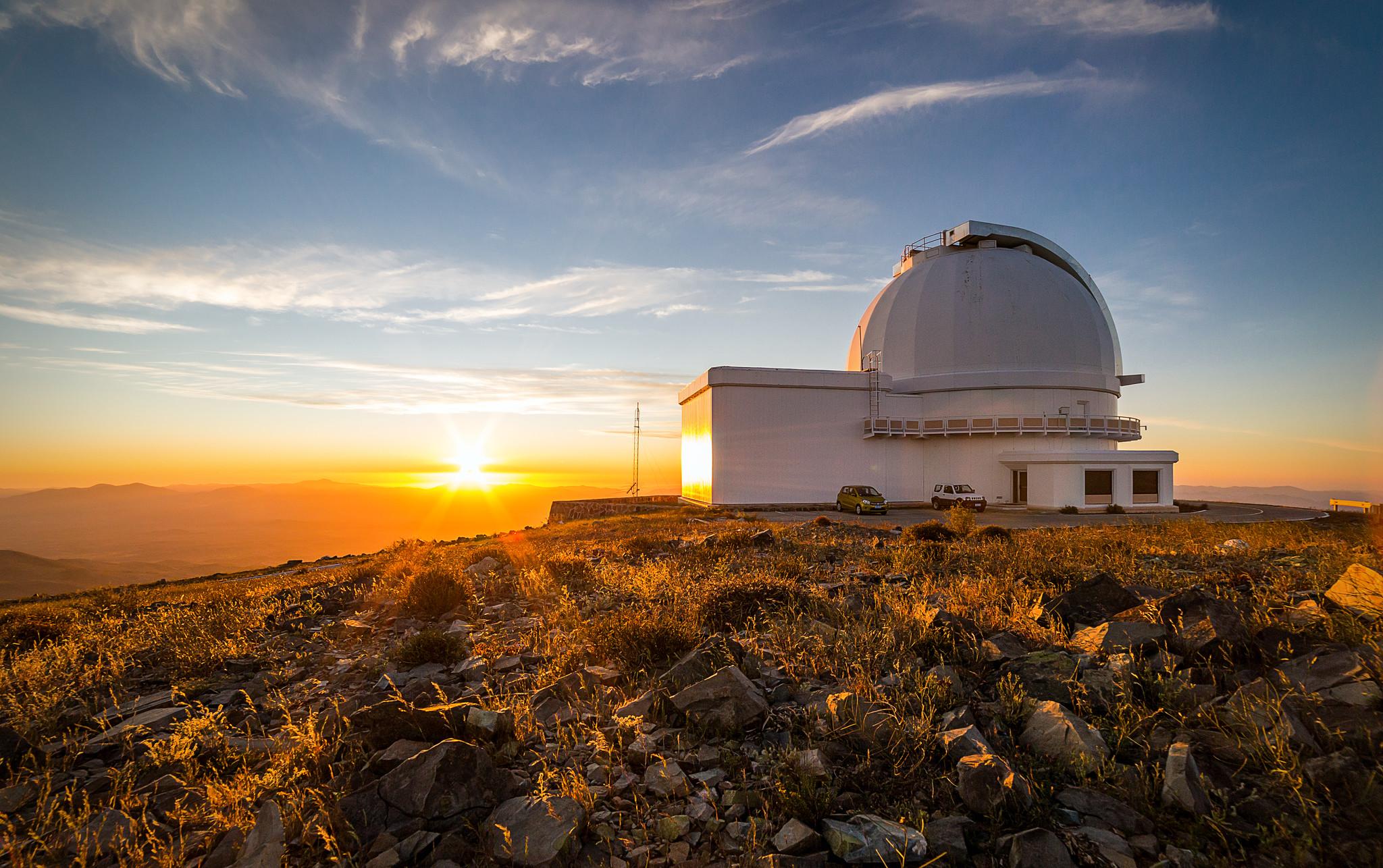 A serene sunset photograph of a white telescope dome on a hill covered with grass and small rocks. To the left of the telescope, the setting Sun tints the landscape in golden shades. A few wispy clouds are scattered in the blue sky above.