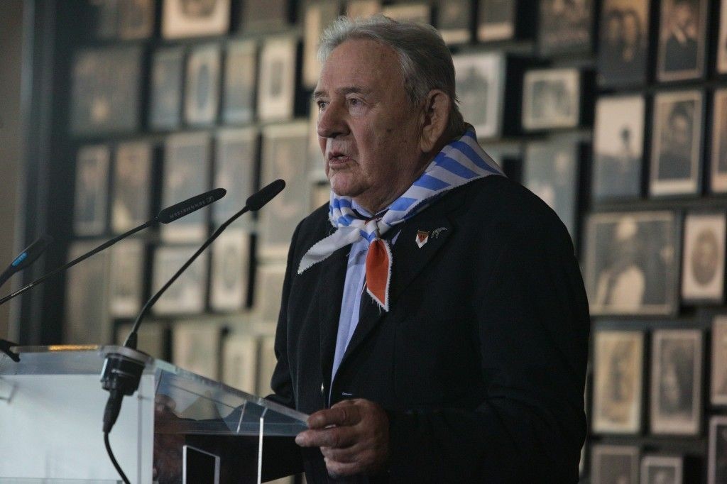 An older man standing behind a lectern with microphones. Around his neck is a scarf with a striped pattern symbolising concentration camp prisoners. Behind him on the wall, a row of blurred photographs showing people. Bogdan Bartnikowski - Auschwitz survivor.