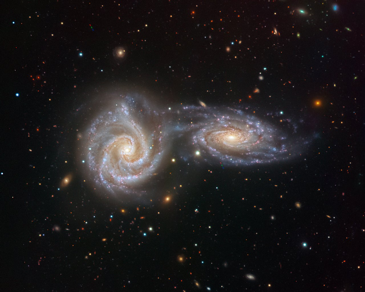 A telescopic image of two spiral galaxies next to each other. The galaxies are nebulous structures with denser arms shaped like spirals that meet at the centre of each galaxy. One galaxy is face-on, and the other somewhat inclined, and they're almost touching. The image is sprinkled with foreground stars and small distant galaxies.