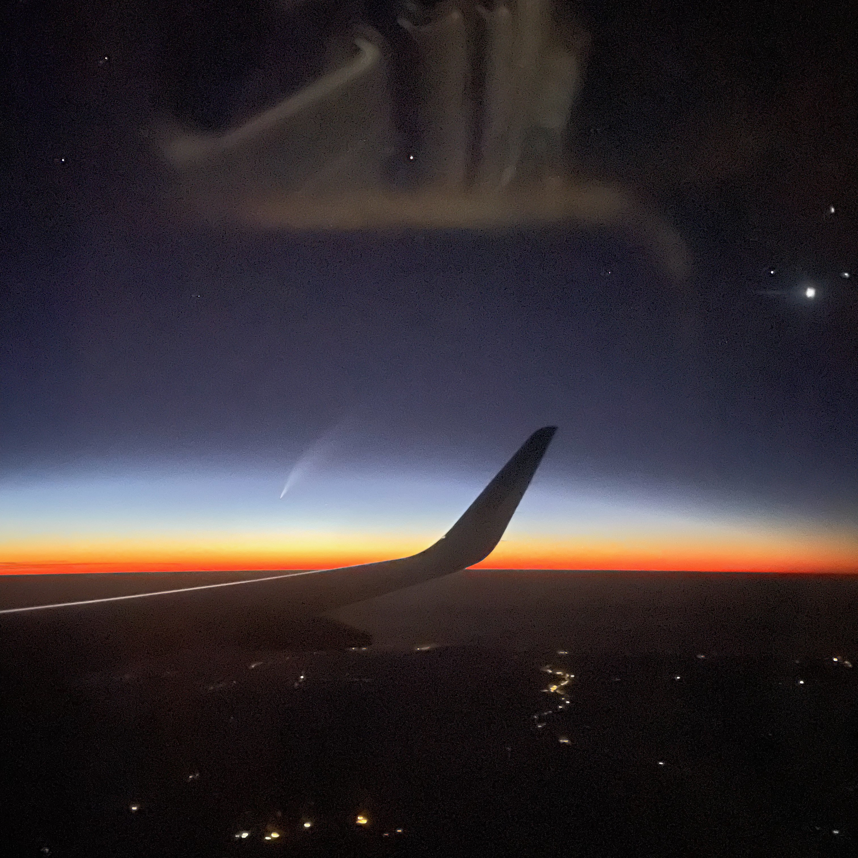 A view from an airplane window during twilight, showing the wing silhouetted against the sky. A comet –a white elongated smudge– hovers above the horizon, surrounded by scattered stars. Below, faint city lights dot the dark landscape.