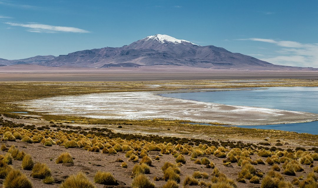 The shore of a turquoise lagoon, sprinkled with white mineral deposits. There's some low vegetation around the shore and, in the background, a dark triangular mountain, its peak covered in snow. The sky is blue and mostly clear, with just a few wispy clouds here and there.