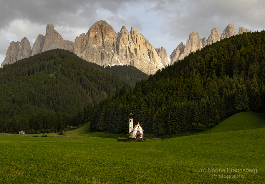 Tiny Church of St. Johann, Val di Funes, sits in a valley in the panorama of the Geisler Mountains here:

https://www.pictorem.com/1939346/Dolomite%20St%20Johann%20Val%20Funes%20Church.html
