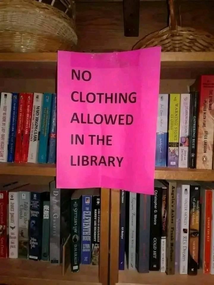 The image depicts a bookshelf filled with various books. A prominent pink sign placed among the books clearly states, "NO CLOTHING ALLOWED IN THE LIBRARY." The surrounding books include a mix of genres, with diverse colors and titles visible, such as "The Pelican Brief," "Effortless," "Pygmalion," and "Kurt Vonnegut." The setting suggests this is a personal library or book collection area where clothing is not permitted.