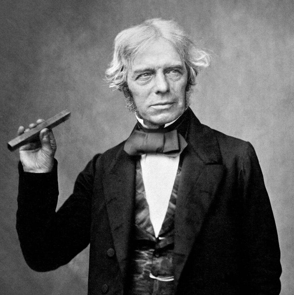 Faraday holding a type of glass bar he used in 1845 to show magnetism affects light in dielectric material.

Michael Faraday. Photographed in the 1850s by Maull and Polyblank as part of their 'Living Celebrities' series. The Faraday volume was published in October 1857.

In this photograph, Faraday is captured with a formal yet dignified demeanor, dressed in traditional Victorian attire—a dark suit with a high-collared white shirt and cravat, reflecting the fashion of the mid-19th century. His face bears a thoughtful expression, with slightly furrowed brows, suggesting the intensity and curiosity of his intellectual life. Faraday's hair is carefully styled, typical of the period, with his receding hairline and sideburns prominently visible. His expression is calm, but his eyes convey sharp intelligence and deep concentration.