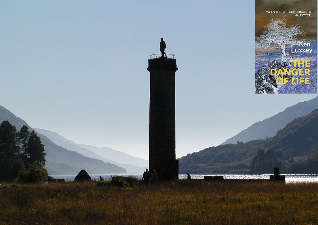 The image shows a view across a field of tall grass to the silhouette of a tall circular column which is topped off by a statue of a man. Beyond the column is a loch which extends away into the distance with wooded mountainsides rising on both sides, distantly lost in mist. The sky is light blue. The front cover of ‘The Danger of Life’ is shown in the top right corner.