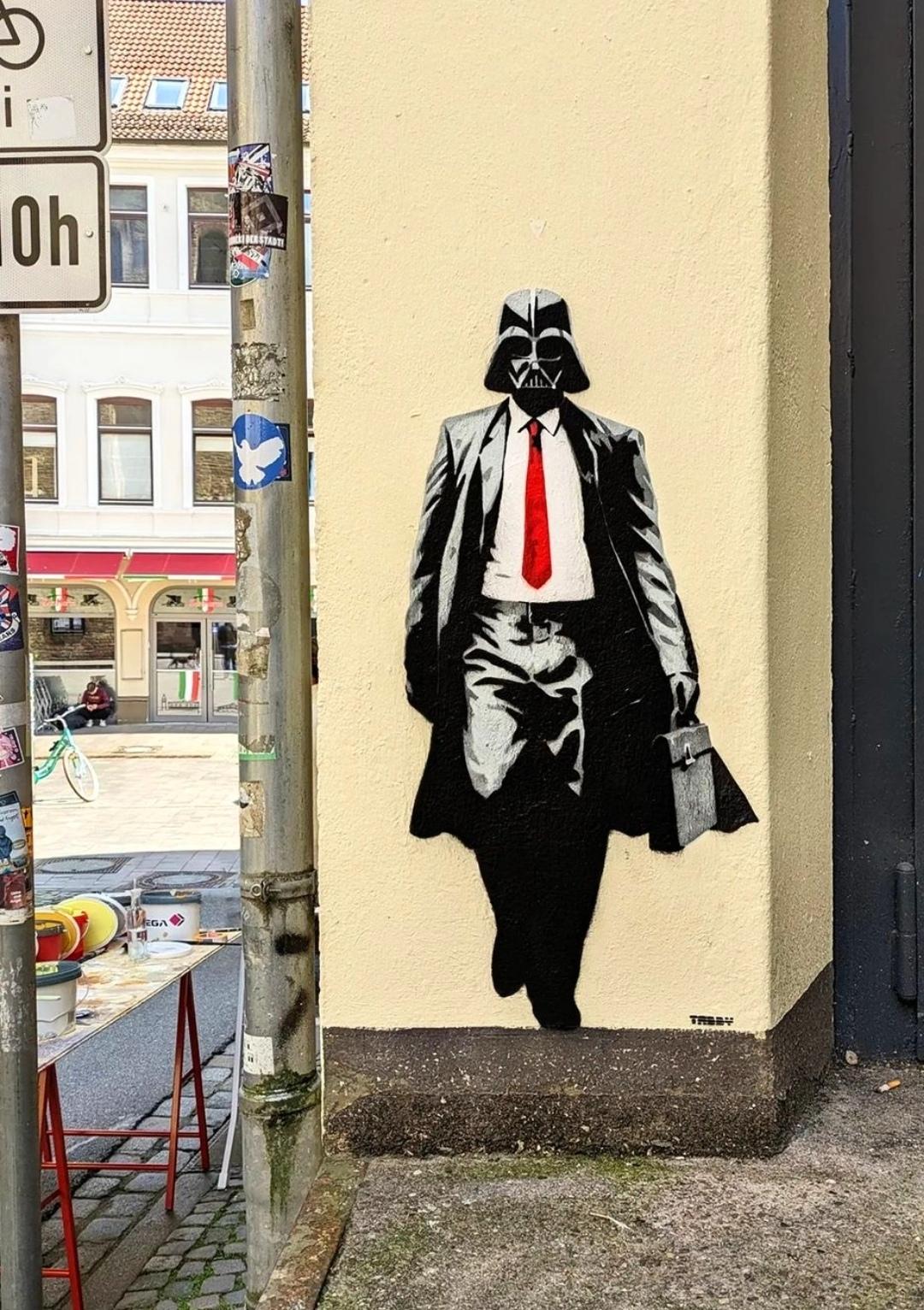 Streetartwall. The mural of a walking Darth Vader (from Star Wars) was sprayed on a narrow exterior wall in the city center next to a black iron door. Darth Vader is sprayed in shades of gray and black using a stencil technique. He is wearing a black business suit, a white shirt, a red tie and a long black coat over it. His typical mechanical black head mask is of course also a must. A very cool and casual mural in an unexpected place in the city.
Info: Darth Vader is the villain from the Star Wars saga. A man who wears black steel armor, a cape and a square, hermetically sealed helmet that supplies him with oxygen.