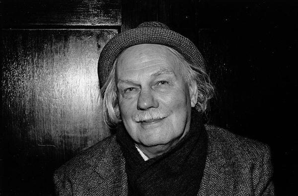 A black-and-white photo of an older Hamish Henderson. He has white mid-length hair, and wears a tweed hat, coat and scarf. He is standing against a wooden door. The photo is from Tobar an Dulchais: https://www.tobarandualchais.co.uk/major-contributors/hamish-henderson?l=en