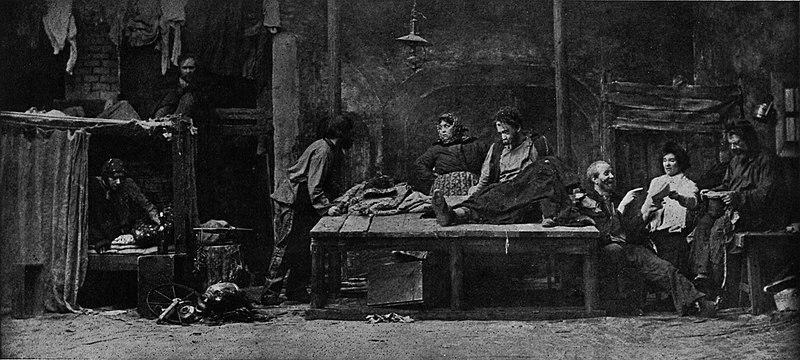 Scene from Act 1 of The Lower Depths, Moscow Art Theater, 1902.

The stage is designed to resemble a cramped, dimly lit basement shelter. The setting emphasizes the play's themes of poverty, despair, and societal neglect.

The backdrop includes rough, deteriorating walls and sparse furnishings, creating a stark and oppressive atmosphere. There are tattered curtains, exposed brickwork, and a hanging lamp, contributing to the sense of deprivation.

The image features a group of impoverished individuals, each distinct in pose and demeanor, reflecting their personalities and struggles.