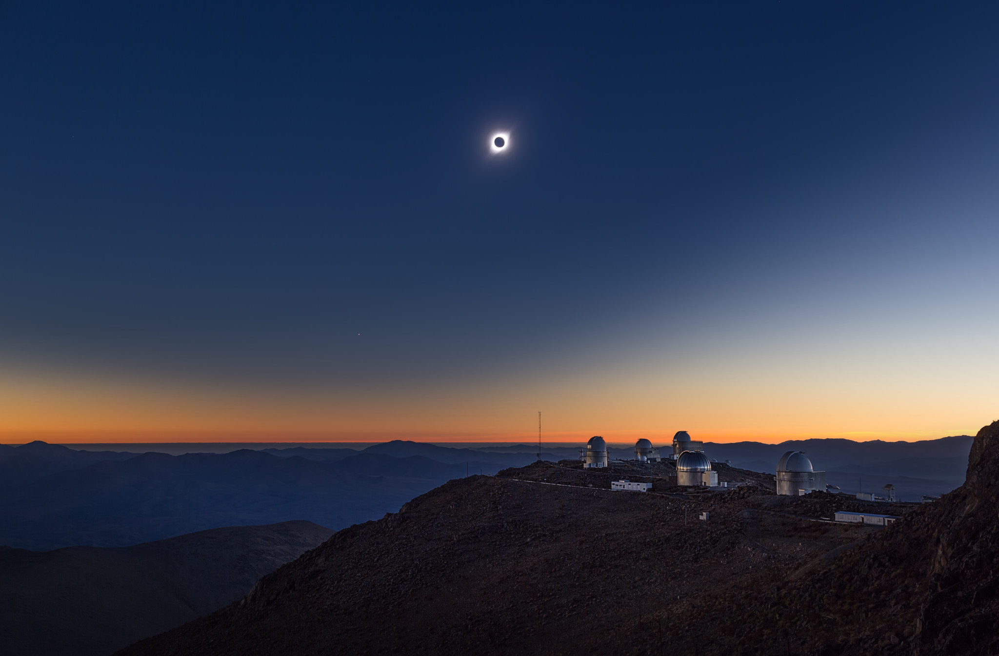 A serene, rocky desert landscape with some round, shiny telescope domes perched on one hill. The sky is completely cloudless, and its colour smoothly changes from golden in the horizon to a deep dark blue higher up. In the middle of the sky, a dark circle is surrounded by a hazy bright glow.