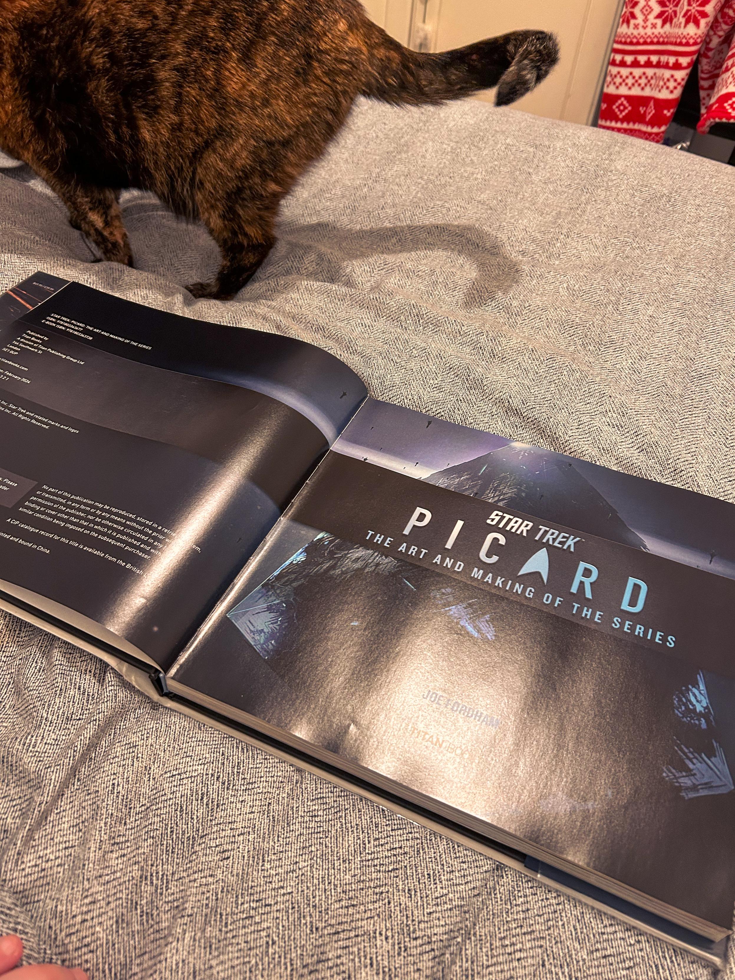 A hardcover book titled Star Trek: Picard - The Art and Making of the Series is open on a bed with a grey herringbone-patterned blanket. The left page contains text, while the right page features the book’s title and design. A tortoiseshell cat is walking across the bed, partially visible from behind.