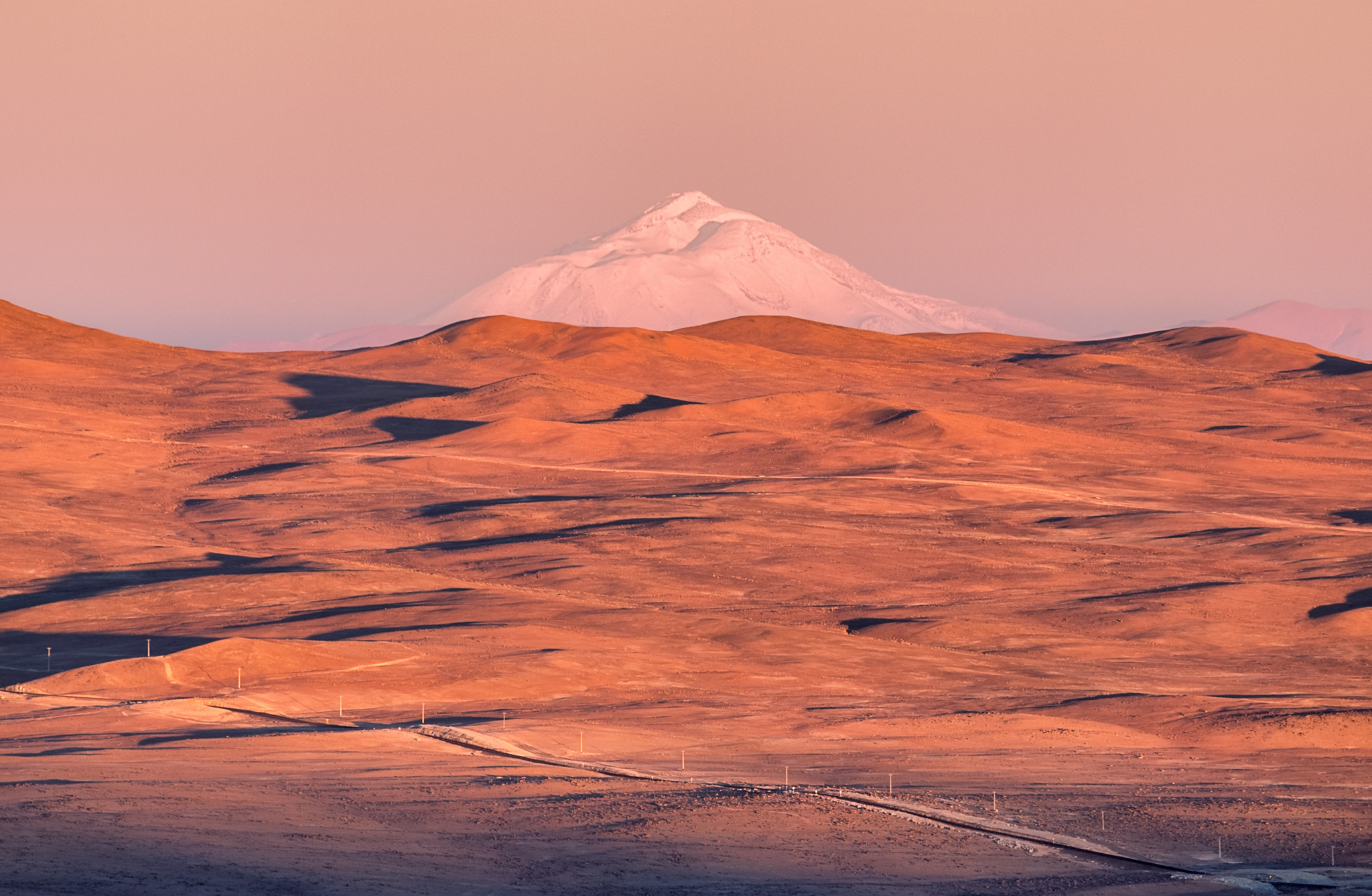 A white, triangular-shaped volcano emerges behind the horizon of a wide expanse of brown undulating hills. The landscape is largely barren, save for a road and a power line seen at the foreground.