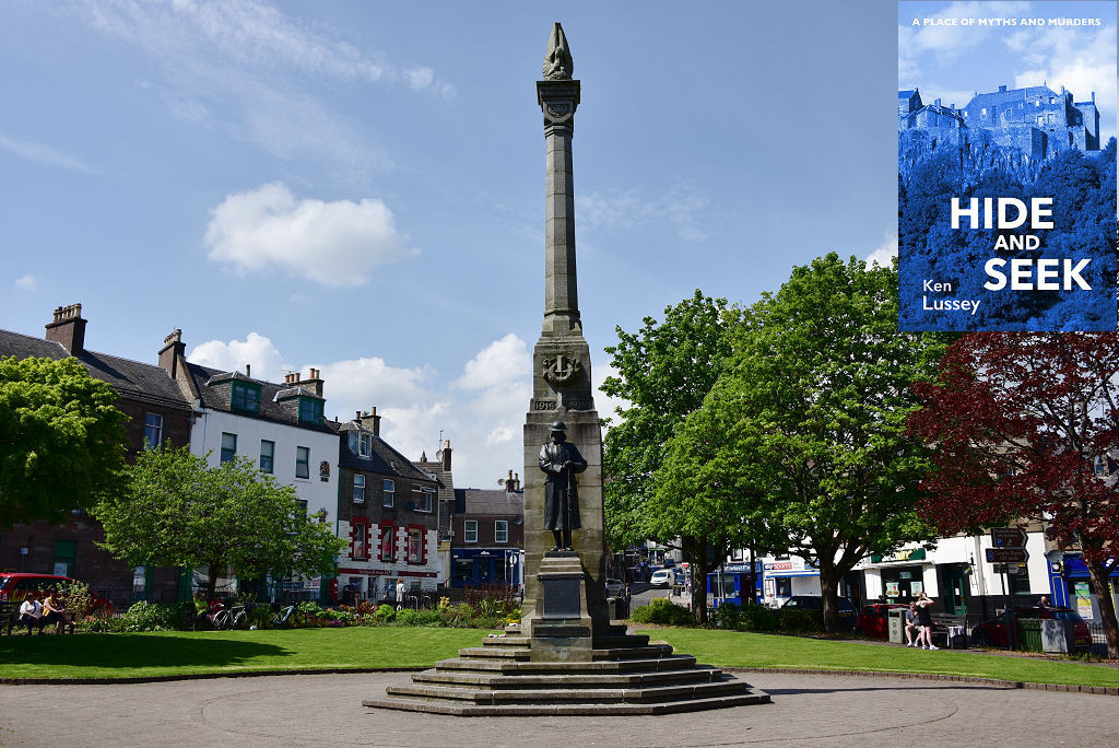 This modern image shows Wellmeadow in the centre of Blairgowrie in Perthshire. In the centre of the image is the tall war memorial with a paved area this side of it and grass beyond, with trees on two sides. The scene is in sunlight. The front cover of ‘Hide and Seek’ is shown in the top right corner.