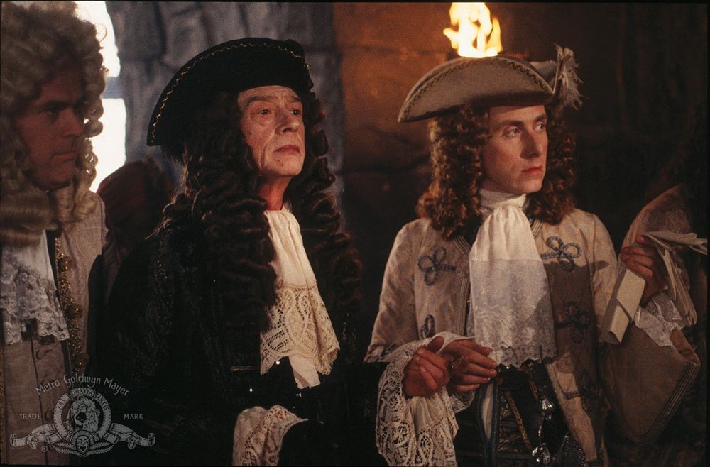 A still from the 1995 film Rob Roy. John Hurt, left, plays the Marquis of Montrose; Tim Roth, right, plays Archibald Cunningham. Both men are dressed in elaborate seventeenth-century costume, with tricorn hats, long curled wigs, lace cuffs and cravats. Hurt's costume, and wig, is dark; Roth's is pale, with contrasting frogging. Both men look intently to the right.