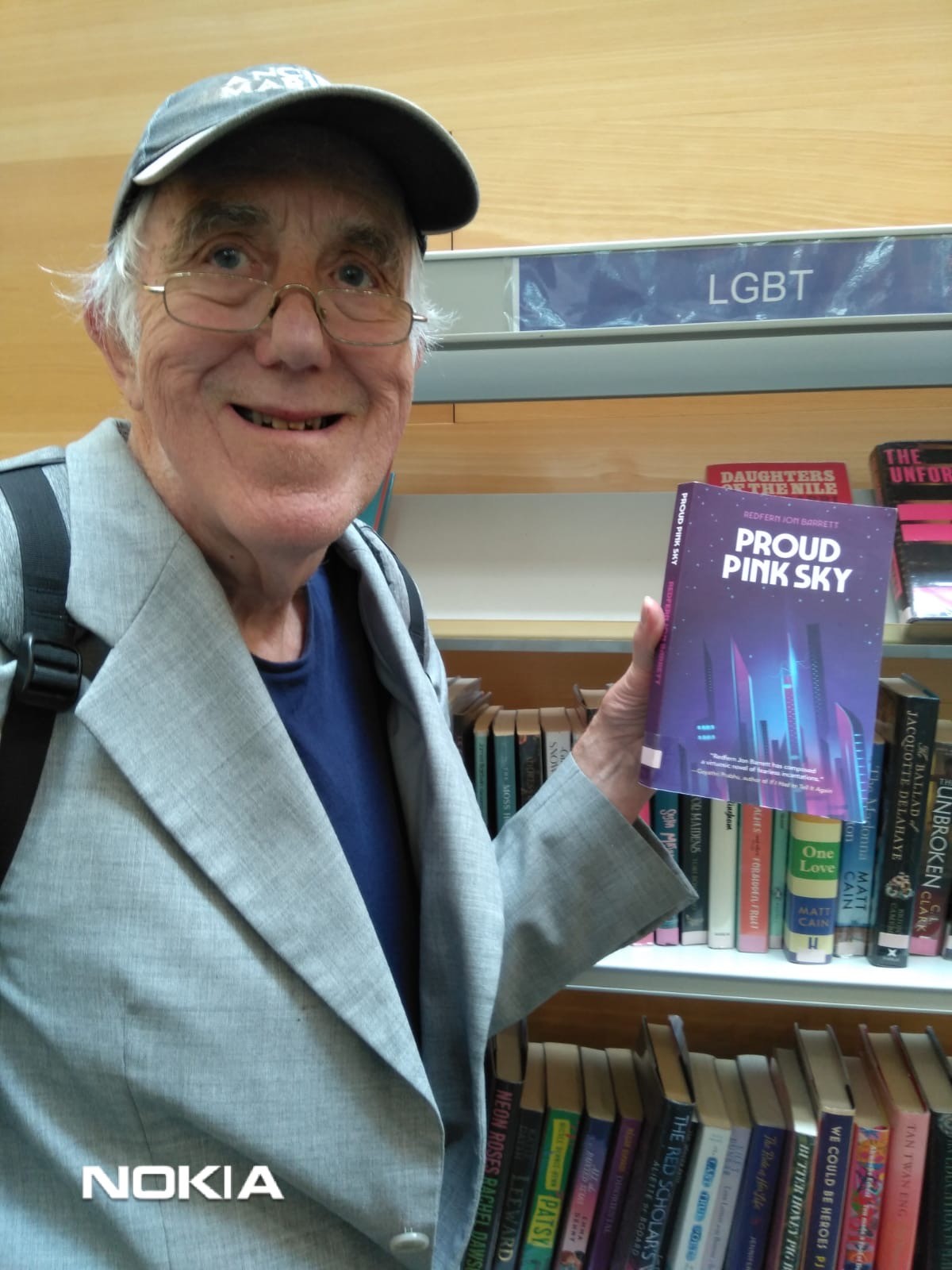 Redfern’s dad standing in front of a library bookshelf, holding up the novel Proud Pink Sky.