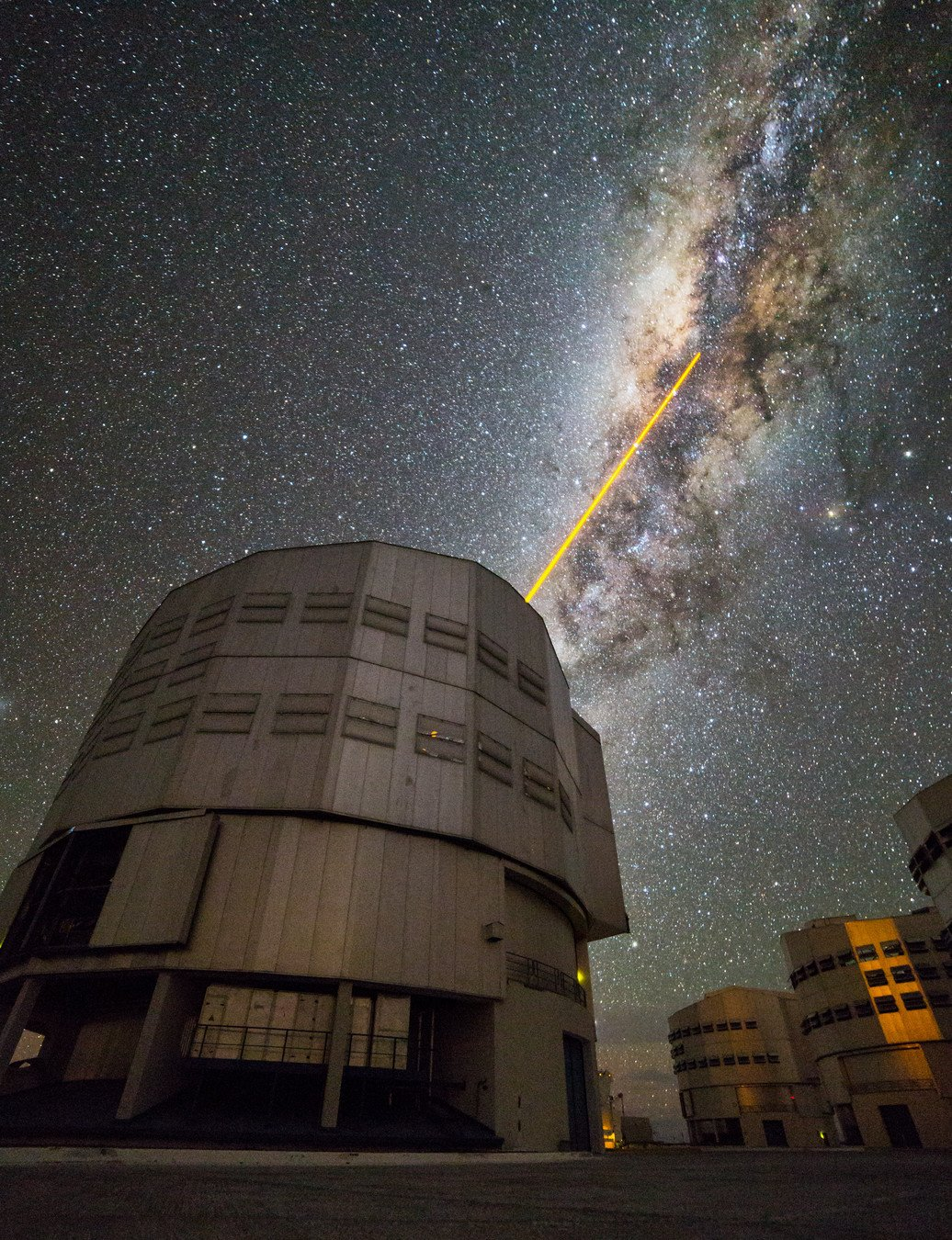 A long-exposure night shot taken at an astronomical observatory under a starry sky. The are several huge cylindrical grey domes. The one in the foreground is shooting a yellow laser up to the sky. The laser is aimed at the centre of the Milky Way, which appears as a bright, somewhat orange band stretching diagonally across the sky, and divided longitudinally by dark clouds.