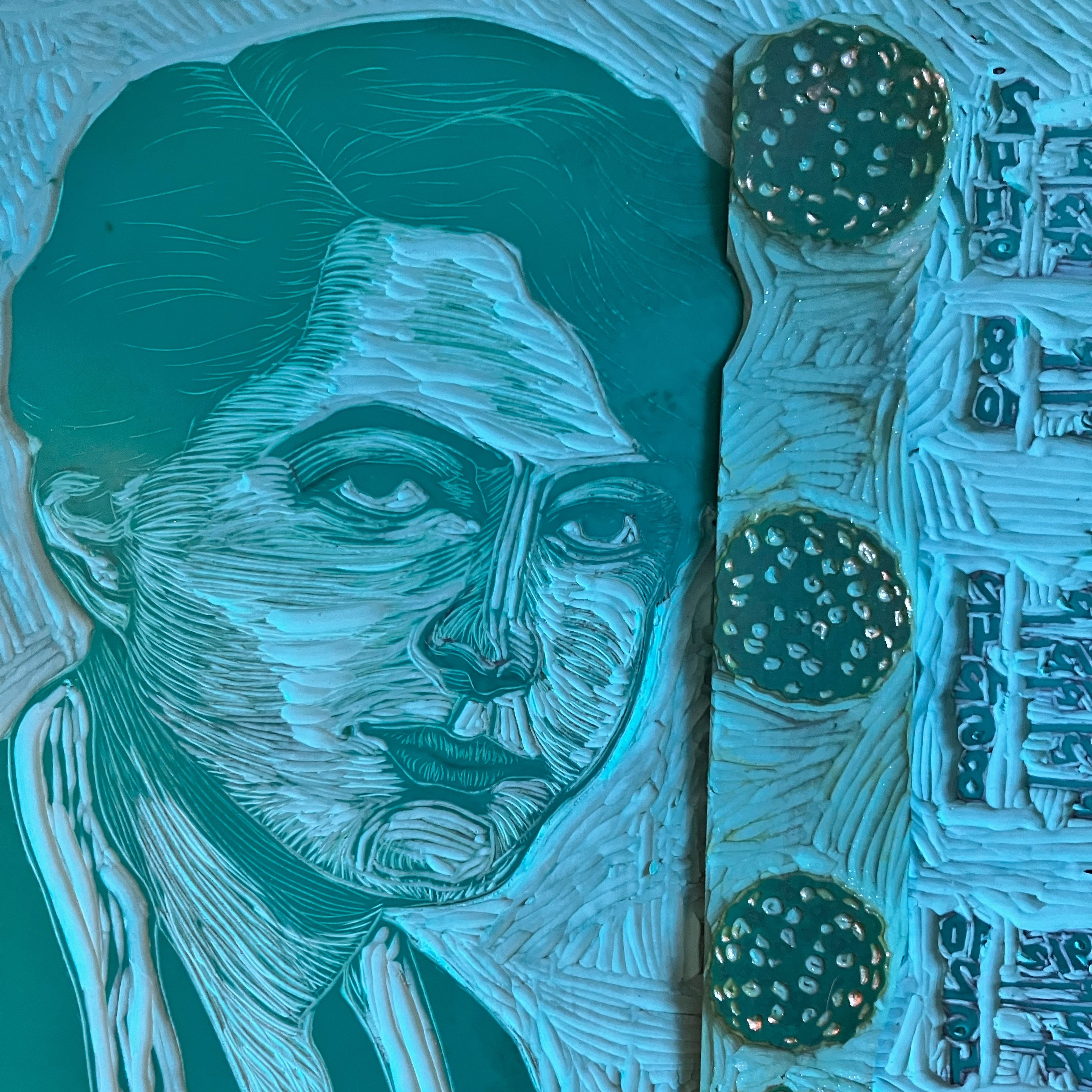 Two green vinyl Lino blocks in progress but mostly carved. On top there’s a narrow one with 3 nuclei which each look like a collection of differing numbers of balls, some carved some not. This is on top of a block with Maria Goeppert Mayer, here as a young, short haired woman with dark shirt and long carved scarf with 3/4 profile and at the edge there’s a bit of a group of numbers and lines.