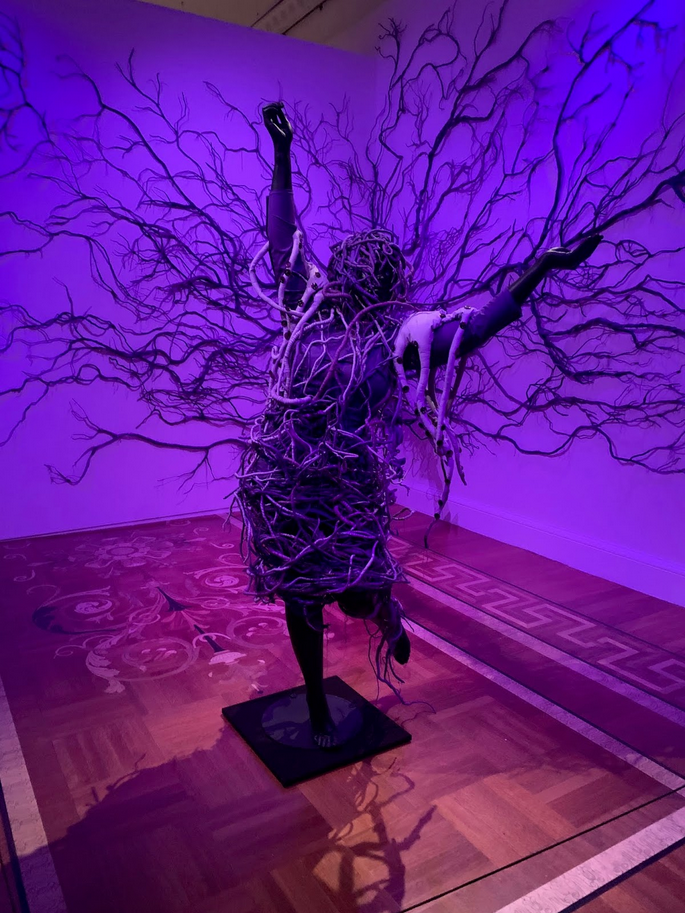 an art installation, a dark figure with arms outstretched, standing up on one leg, it is purple with purple walls and dark purple roots enveloping and covering her