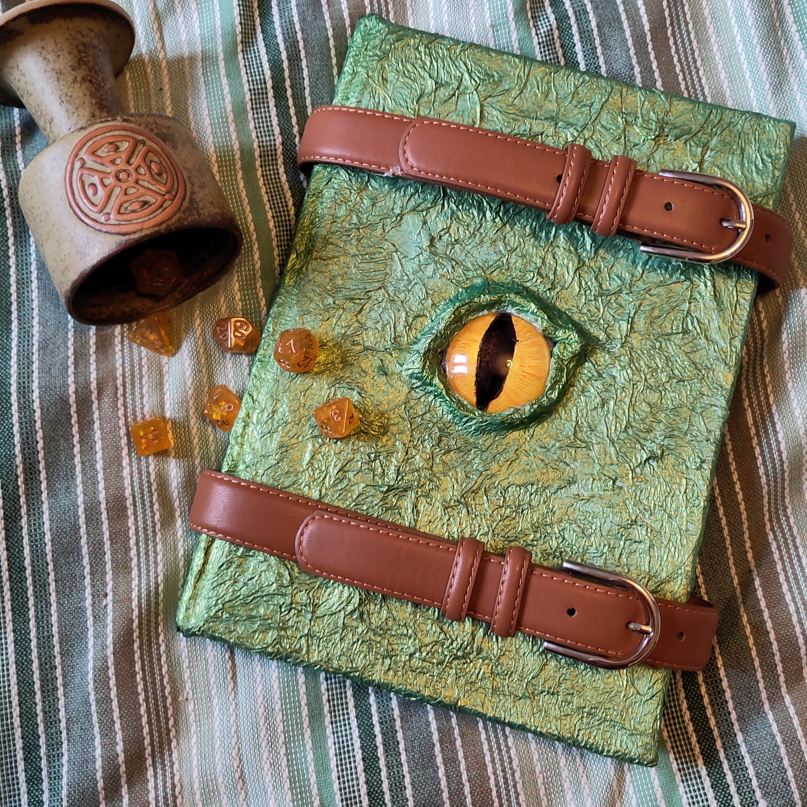 The journal is an A4 notebook that has been covered in pva coated kitchen roll sculpted to look like dragon skin and painted with two tone iridescent paint that captures different shades of green from different angles. In the centre it has been sculpted to make raised eyelids, and a yellow dragon eye set in it with a slit iris like a lizard - this was painted on the back of a glass lens. It is bound together with two leather belts. Beside it is a goblet pouring out DnD dice