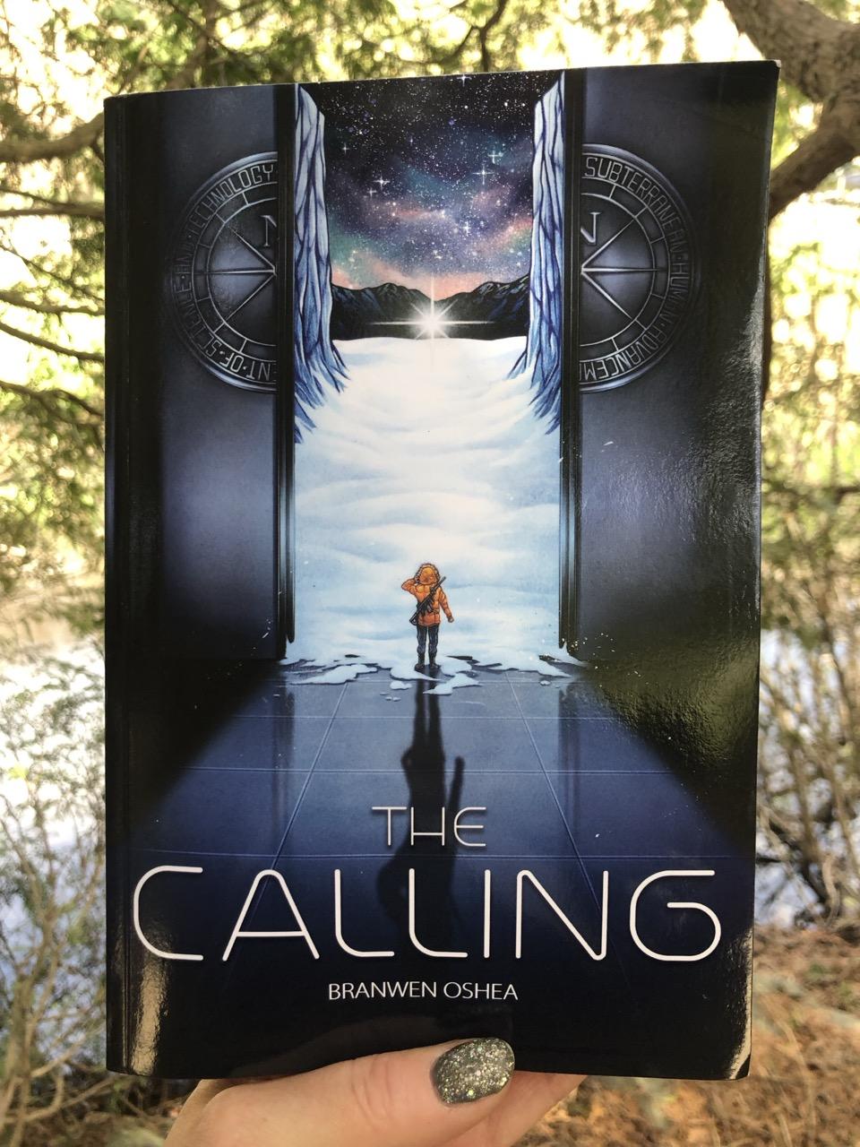 Author’s hand holding a paperback of The Calling outside. The cover features a teen exiting a massive elevator into a nighttime Arctic landscape. Far in the distance, on the snow, sits a strange star.