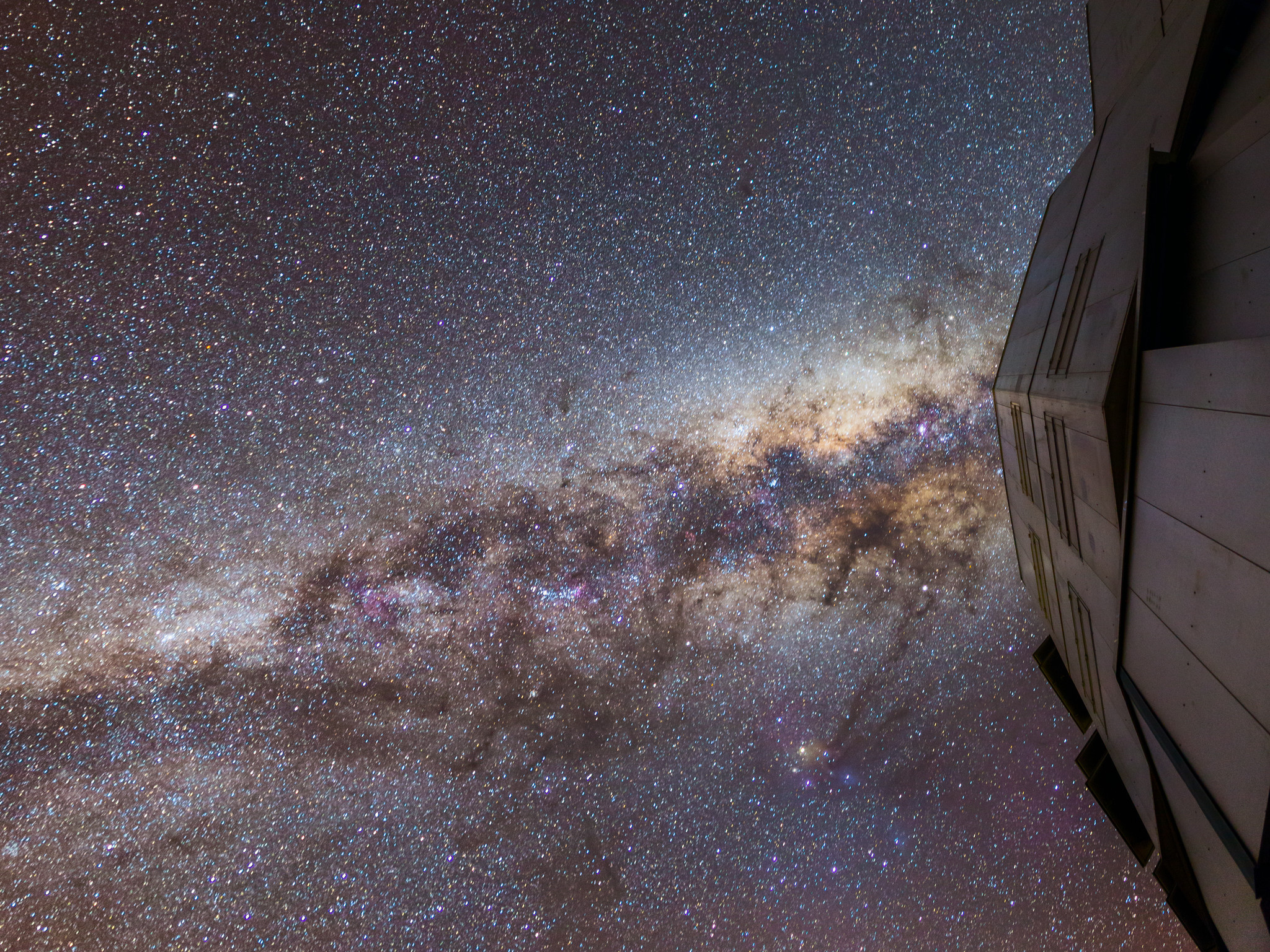 A long-exposure image of the night sky, looking directly overhead. The sky is sprinkled with a myriad of stars. There's a dense band of stars –the Milky Way– crossing the image horizontally, getting brighter and denser towards the right. This band is interrupted here and there by dark wispy dust lanes. To the right of the image there's a grey and very slightly reflective metallic wall with angular corners. The camera is pointed up right next to this wall.