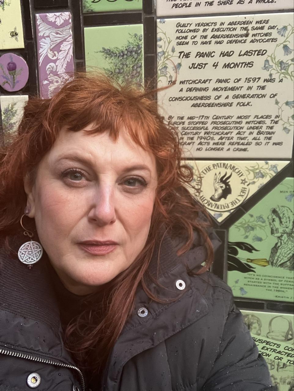 A red haired middle aged white woman squinting and frowning next to a tiled wall of botanical illustrations and quotes that look like xeroxed zines. The tiles are green, beige and pink. The one next to her face says “Hex the Patriarchy” and has a hand creating a shadow-devil shape. 