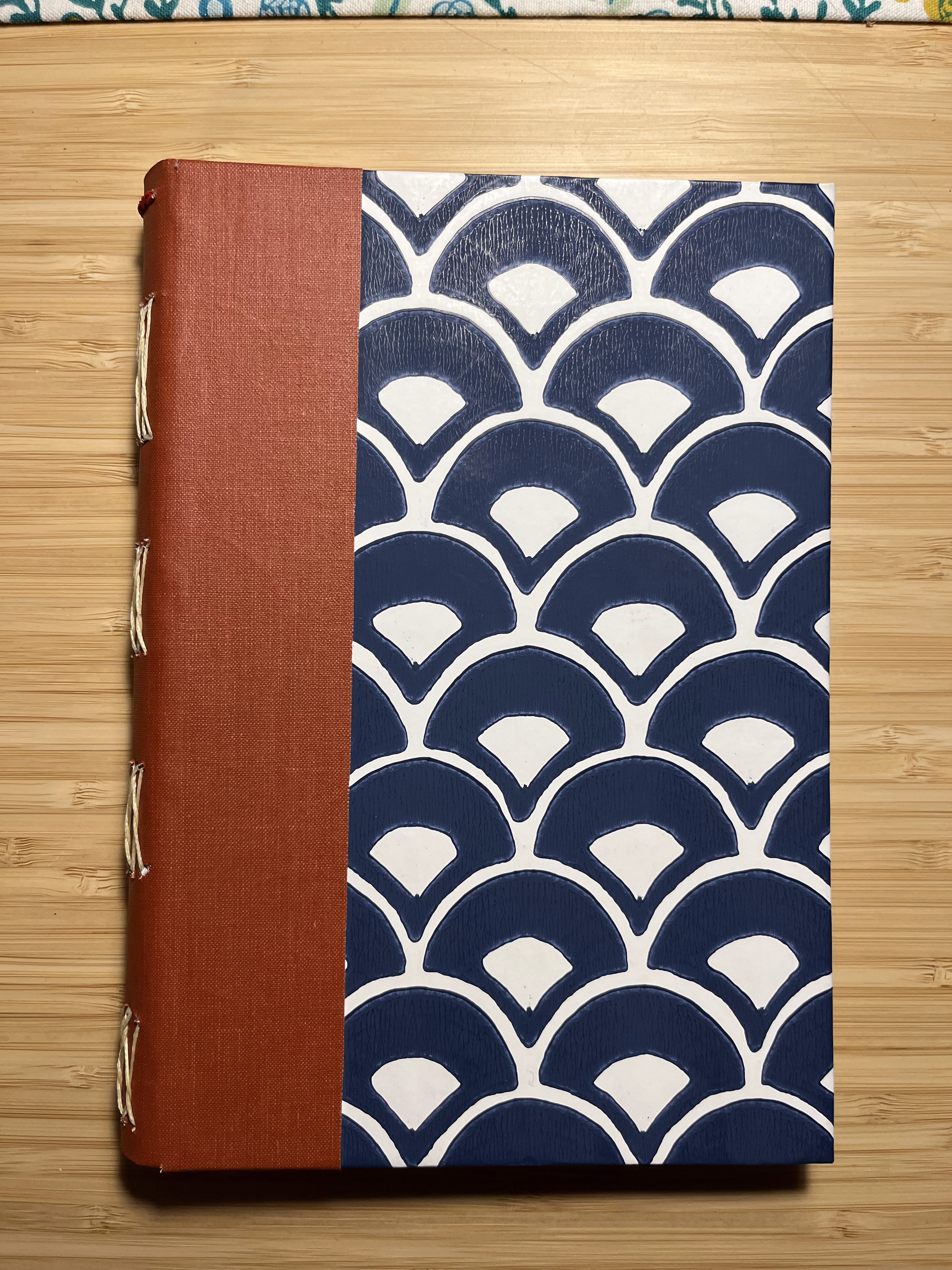 Closed handmade book with a brown spine and blue-and-white patterned cover on a bamboo desk.