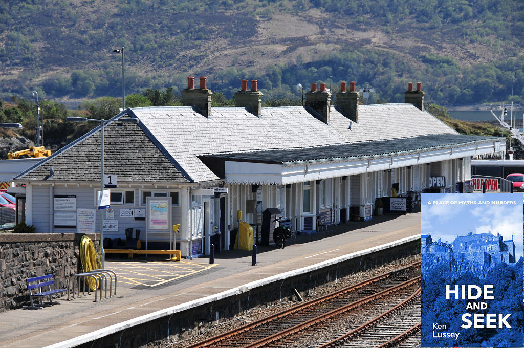 This modern image shows Kyle of Lochalsh railway station. There are tracks in the foreground leading to the right and the station is made of wood with a canopy and a slate roof. There’s a glimpse of a loch in the background and beyond it is a wooded hillside. The scene is in sunlight. The front cover of ‘Hide and Seek’ is shown in the bottom right corner.