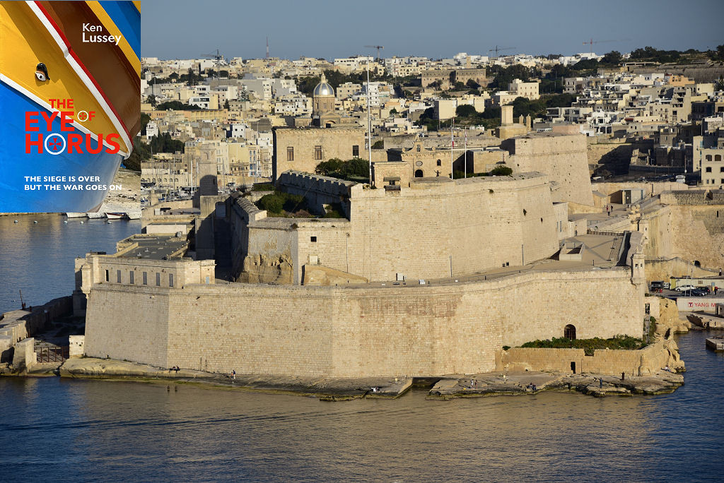 The image shows a telephoto view of Fort St Angelo seen slightly from above and across Malta’s Grand Harbour. The high walls of the fort rise in two layers of bright light stone from the waters of the harbour, which wrap around both sides of it. There are buildings in the upper parts of the fort and beyond it is Birgu. The scene is in bright sunlight which is coming slightly from the right. The front cover of ‘The Eye of Horus’ is shown in the top left corner.