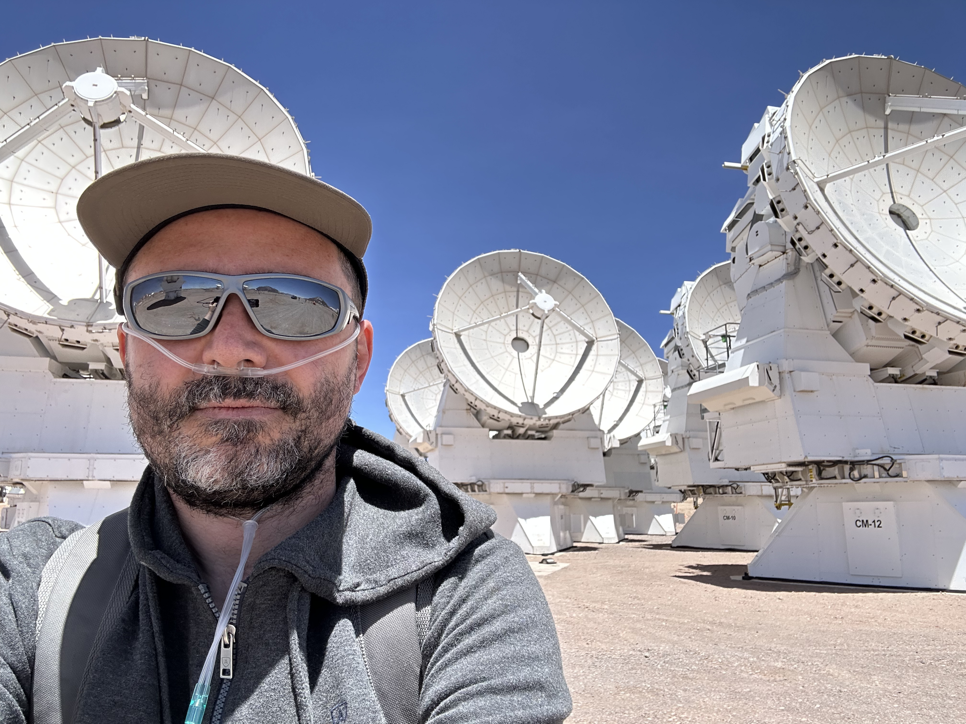 A selfie of a man wearing sunglasses, a cap and a thin respiratory tube attached to his nostrils. In the background there are several huge, white antennas facing the camera, under a clear blue sky.