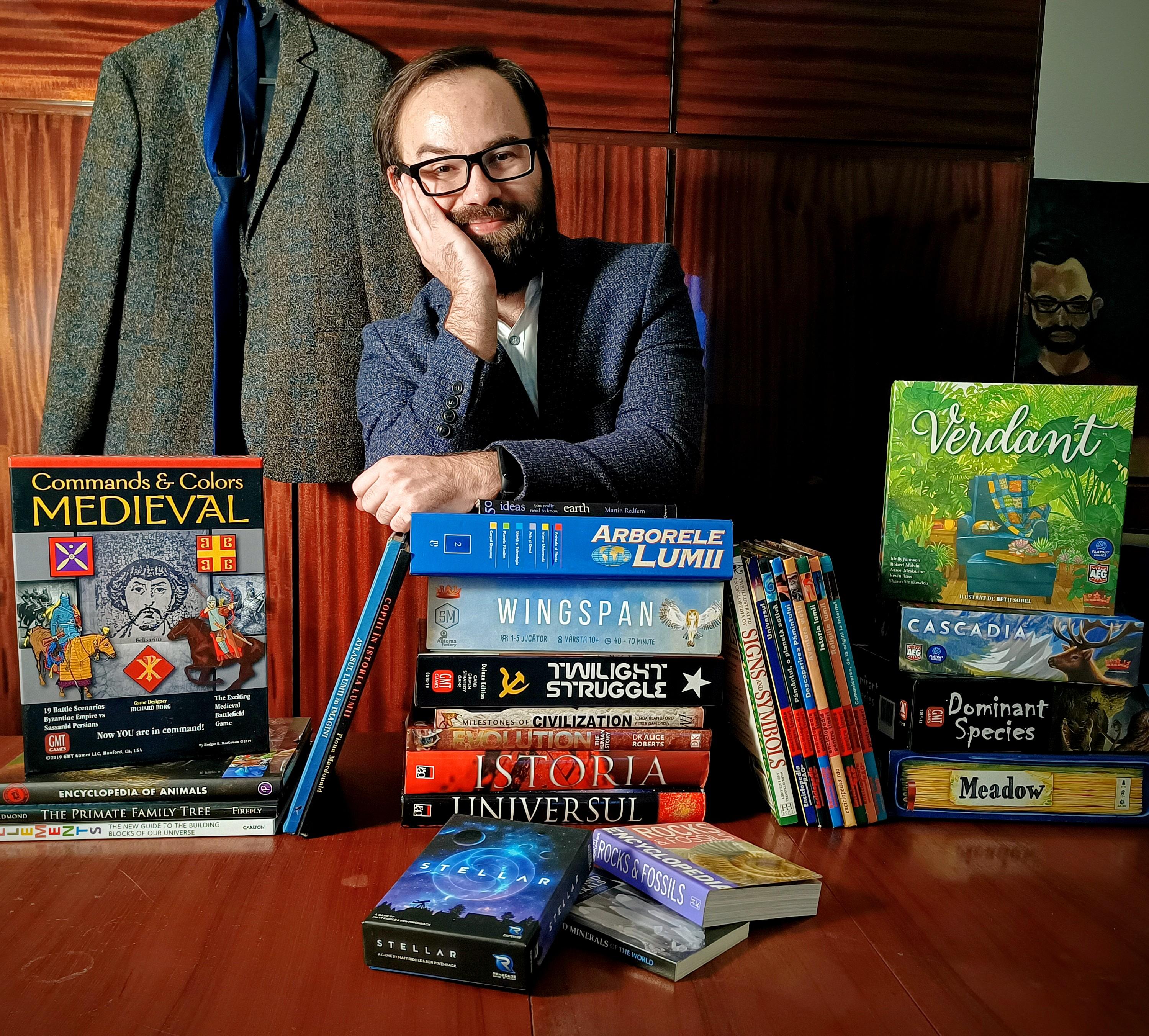 A male person with glasses and beard watching at the camera, leaning over a bunch of books and board games.