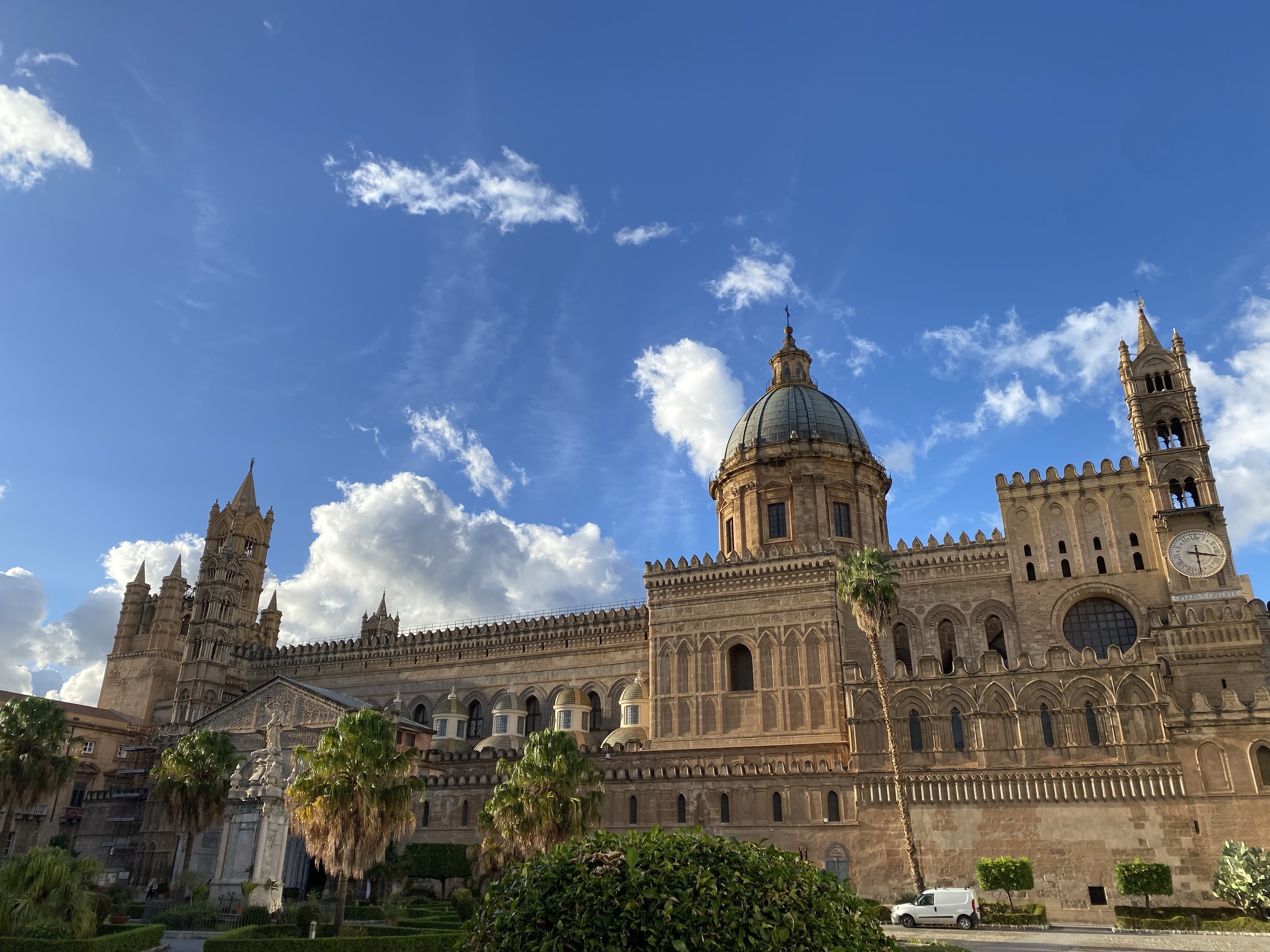 Cathedral with ornate architecture under a blue sky with clouds. On the right can be seen a clock tower and a large spire dominates the middle. Some green scenery surrounds the place of worship.