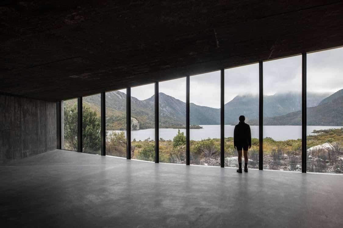 A view of cradle mountain from a row of floor to ceiling windows. The interior is very dark, letting the scene before it dominate. The view has rocks and shrubs in foreground. A lake in the mid and many peaks behind. Some shrouded in fog. 