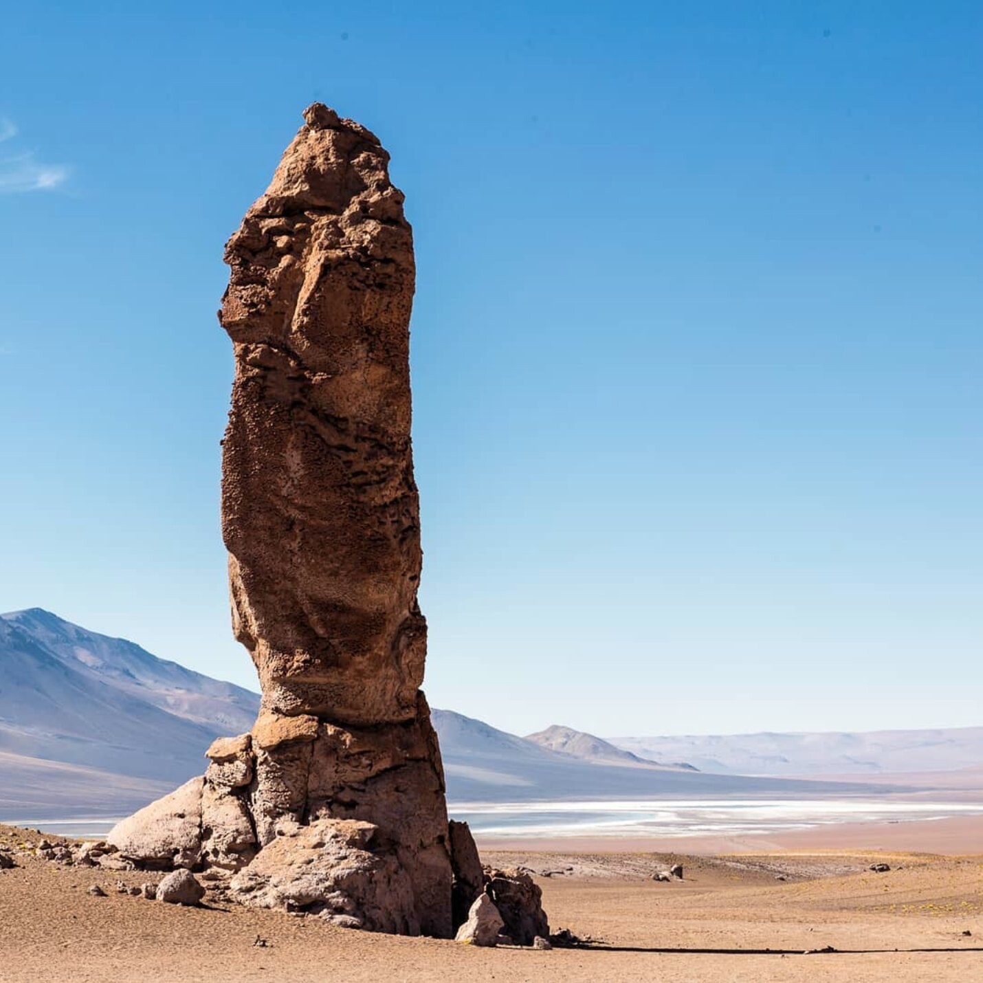 A tall and thin rocky structure, brown and with a roughly cylindrical shape, rises above the ground in an otherwise flattish desert landscape. In the background there's a blue and white salt flat, and some mountains in the distance, under a clear blue sky.