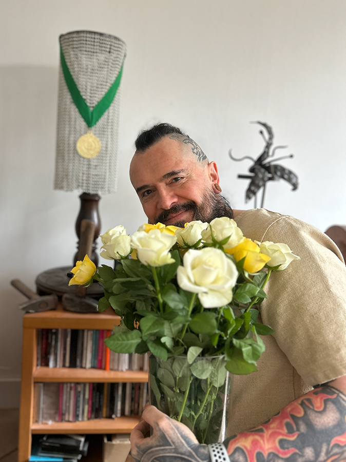 Bjørn with a bucket of roses. A gold medal for Best Historical Fiction Novel hangs in the background. There are a few blacksmithing hammers (not staged, I just like having them where I see them) and a forged plant). Bjørn is wearing a linen tunic and he’s very, very happy.