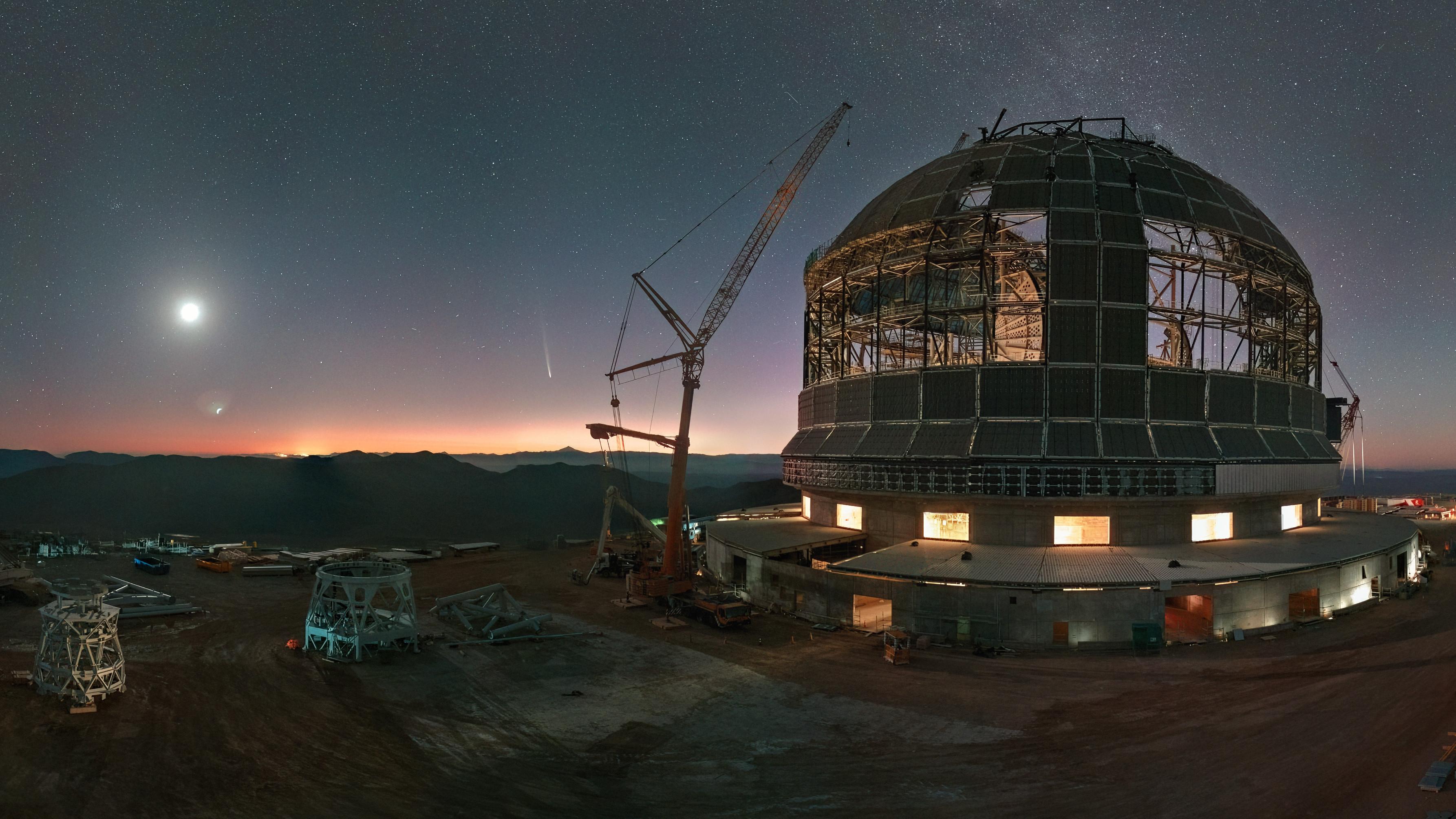 A night shot of a huge metallic dome under a starry sky. The dome is partially covered with rectangular panels, but there are openings revealing complex mechanical structures inside. There's a gigantic crane next to the dome, and some smaller cylindrical structures with criss-cross beams on the ground next to the dome. There's an elongated white smudge of light on the sky –– the comet –– above a golden horizon that signals the imminent sunrise. The moon shines brightly to the left.