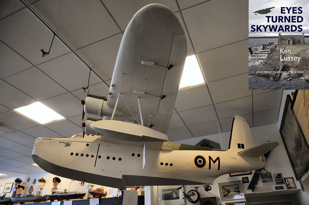 This image is looking up at a large model of an aircraft hanging from a white ceiling with panels. We are looking from the left side of the aircraft, which is a white flying boat with four engines and lots of circular windows in the fuselage. It has an RAF roundel on the rear fuselage with a large letter M behind it. The front cover of ‘Eyes Turned Skywards’ is shown in the top right corner.