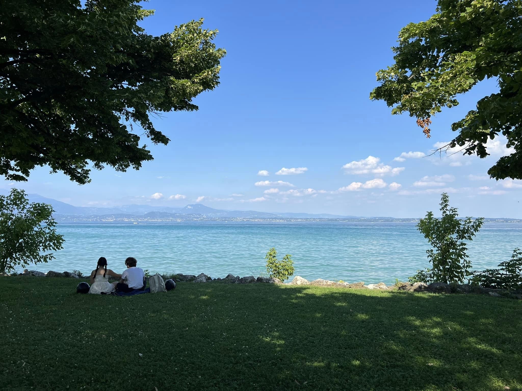 A serene lakeside scene in Sirmione, captioned "Love is in the air." Two people are seated on a grassy area under the shade of large trees, sharing a quiet moment together. They face a tranquil, azure lake with a backdrop of distant mountains under a clear, blue sky dotted with fluffy white clouds. The setting exudes calmness, natural beauty, and a sense of romance.