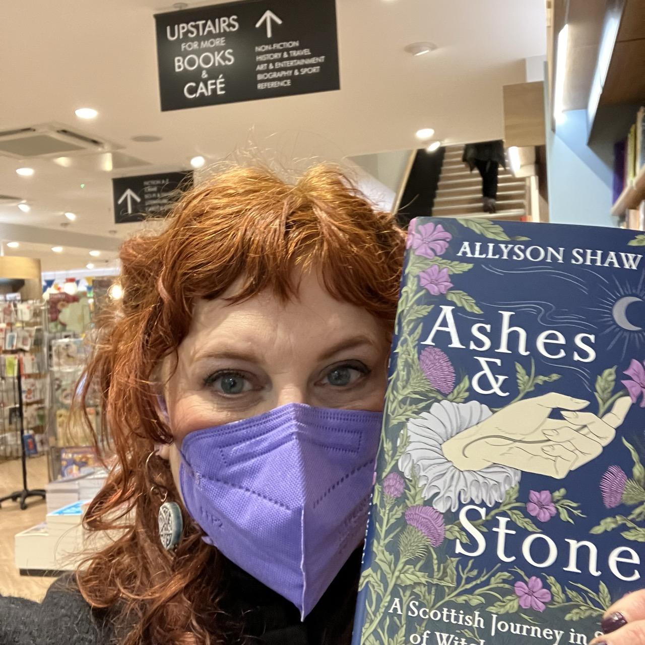 Image of a white, red haired woman in a lavender N95 mask holding a book next to her face. It says “Ashes and Stones” in a white serif font and the book is dark blue with thistle and herb robert flower illustrations. 