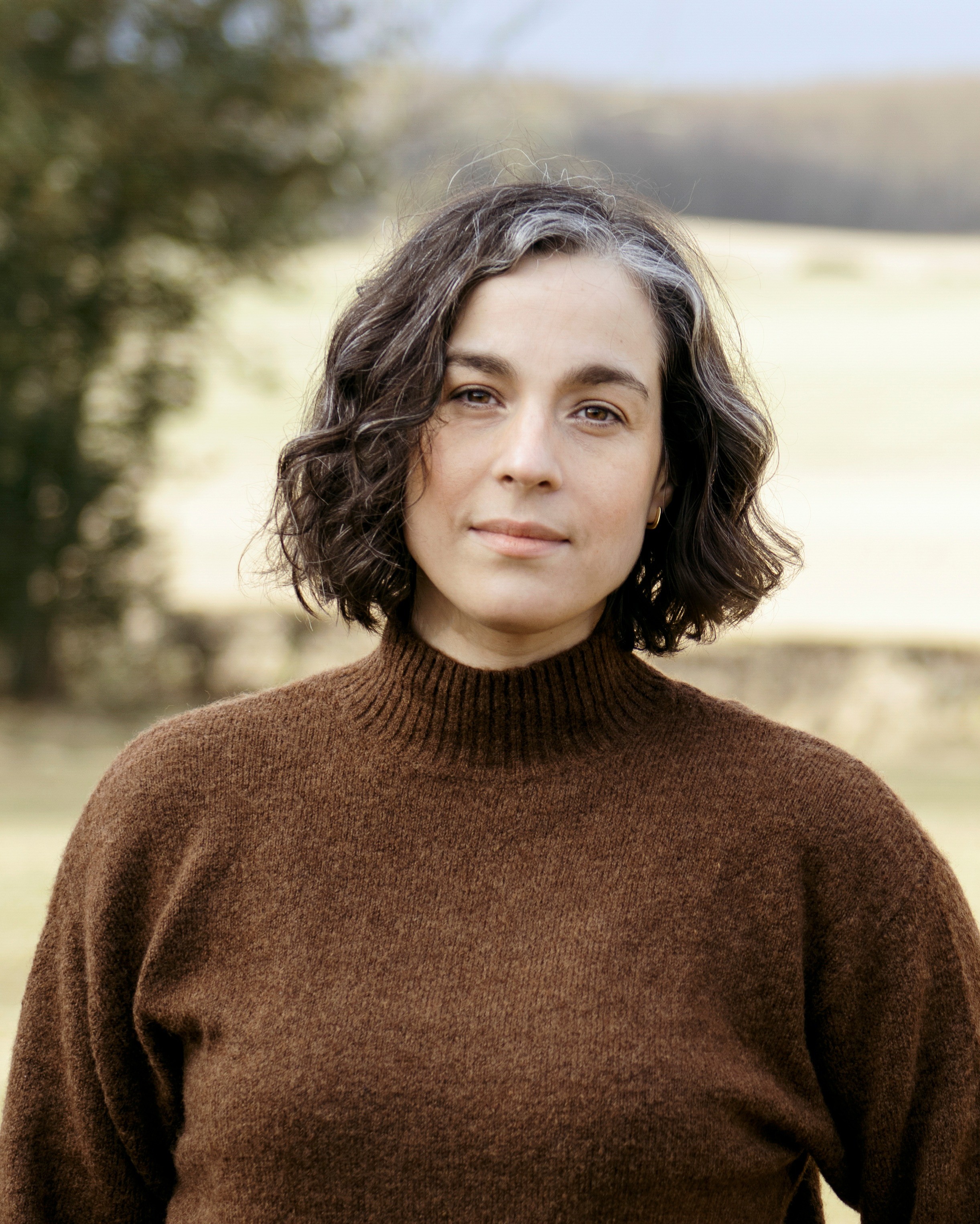 A colour photograph of a middle-aged woman with brown hair, wearing a brown jumper. In the front of her hair are two grey streaks and behind her is fields and trees. She is looking directly at the camera and has a soft smile on her face. 