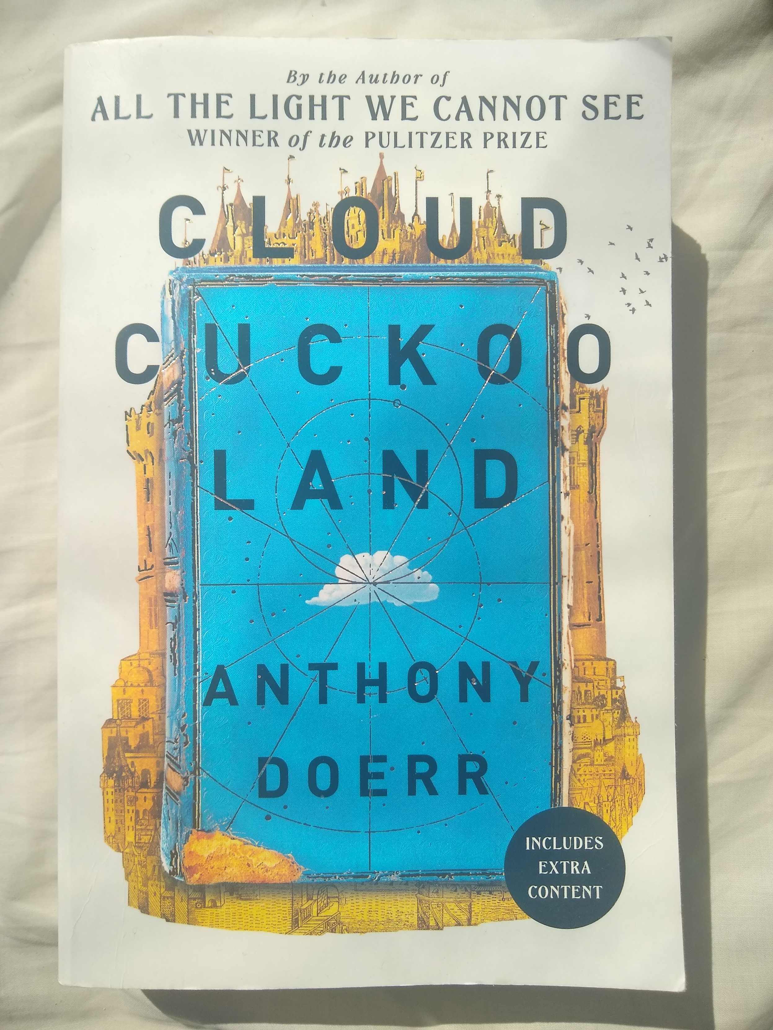 Book cover showing a blue book with a white cloud on the cover, in front of a yellow city with many spires.