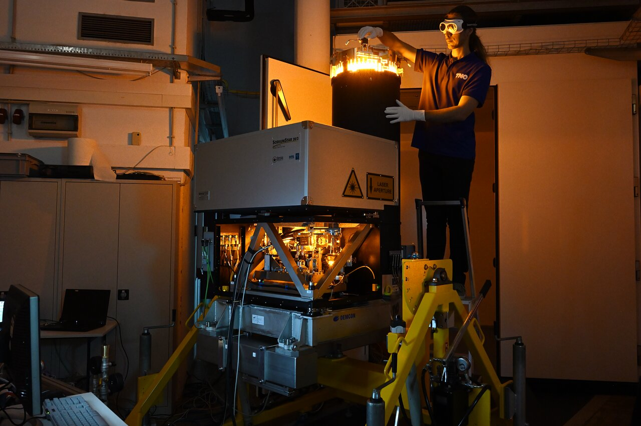 At the centre of the image, a big square machine is standing, its top part a closed grey box, containing a laser, resting on top of an illuminated network of intricate machinery. It is standing on a yellow slider. To the right of the machine, a man wearing safety goggles and gloves is standing on a stepping ladder, installing a part on a big black vertical tube, as high as he is, attached to the back of the square machine. On top, the tube is glowing yellow.