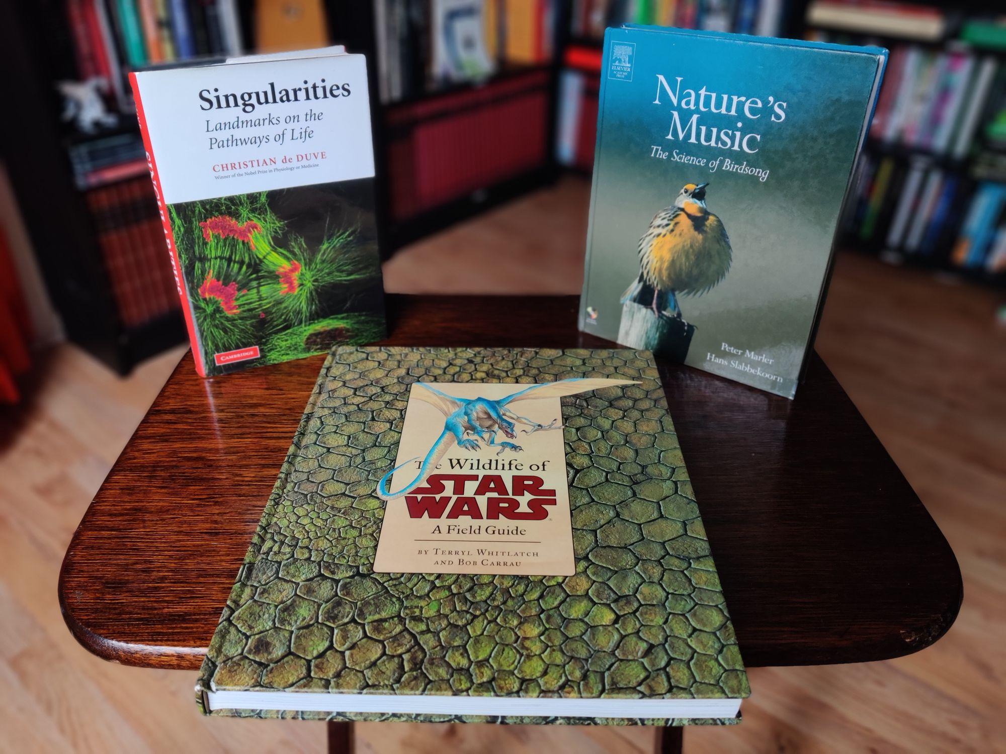 A photo of three books standing on a small, brown, wooden table. On the left, standing up, Singularities: Landmarks on the Pathways of Life, showing a fluorescent microscopy picture with structural elements of cells visible in green and red. In the middle, lying down, The Wildlife of Star Wars, showing a picture of a blue, winged fantasy creature on a background of green, scaly skin. On the right, standing up, Nature's Music: The Science of Bird Song, showing a yellow bird with a puffed-up chest and its beak wide open sitting on a pole. The out-of-focus background shows black shelves full of books and part of the beech-coloured laminate floor.