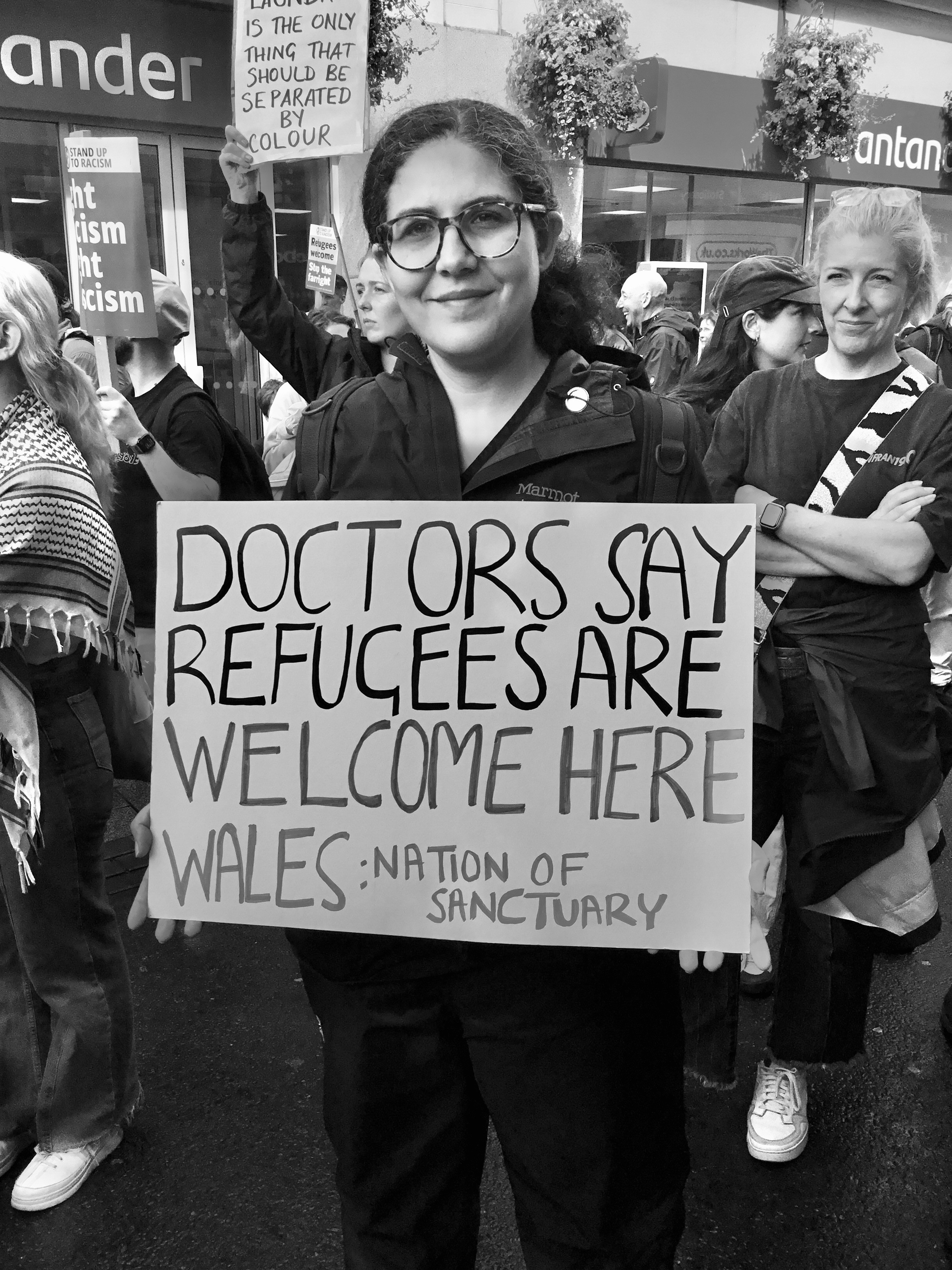 A white person holds a placard saying ” DOCTORS SAY REFUGEES ARE WELCOME HERER WALES: NATION OF SANCTUARY” Black and white image.