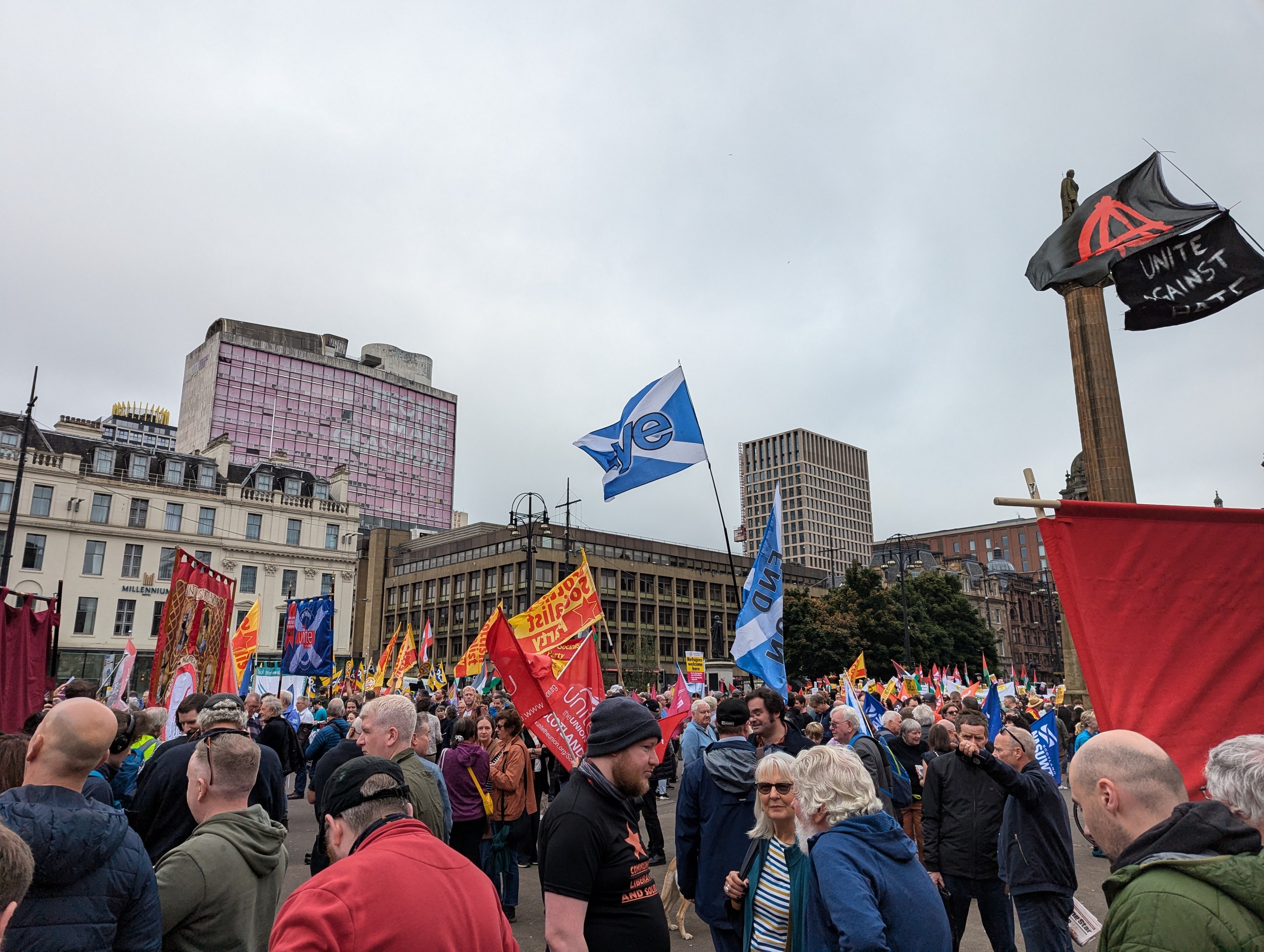 A photo of a crowd of people in Glasgow's George Square at the Stand Up to Racism rally. There are a tonne of flags, signs and banners from different trade unions and campaign groups.