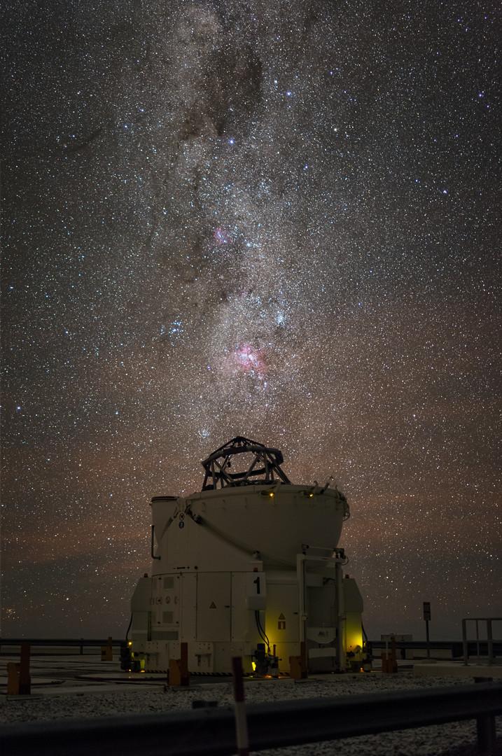 A night photograph of a telescope under the stars. The telescope dome is white and looks like an eggcup, with part of the telescope frame sticking out of the top. The sky is crowded with countless stars. The Milky Way –a dense band of stars– stretches vertically above the telescope, as if the telescope was a chimney with white celestial smoke coming out it. The Milky way is sprinkled with pink nebulae and dark clouds of cosmic dust.