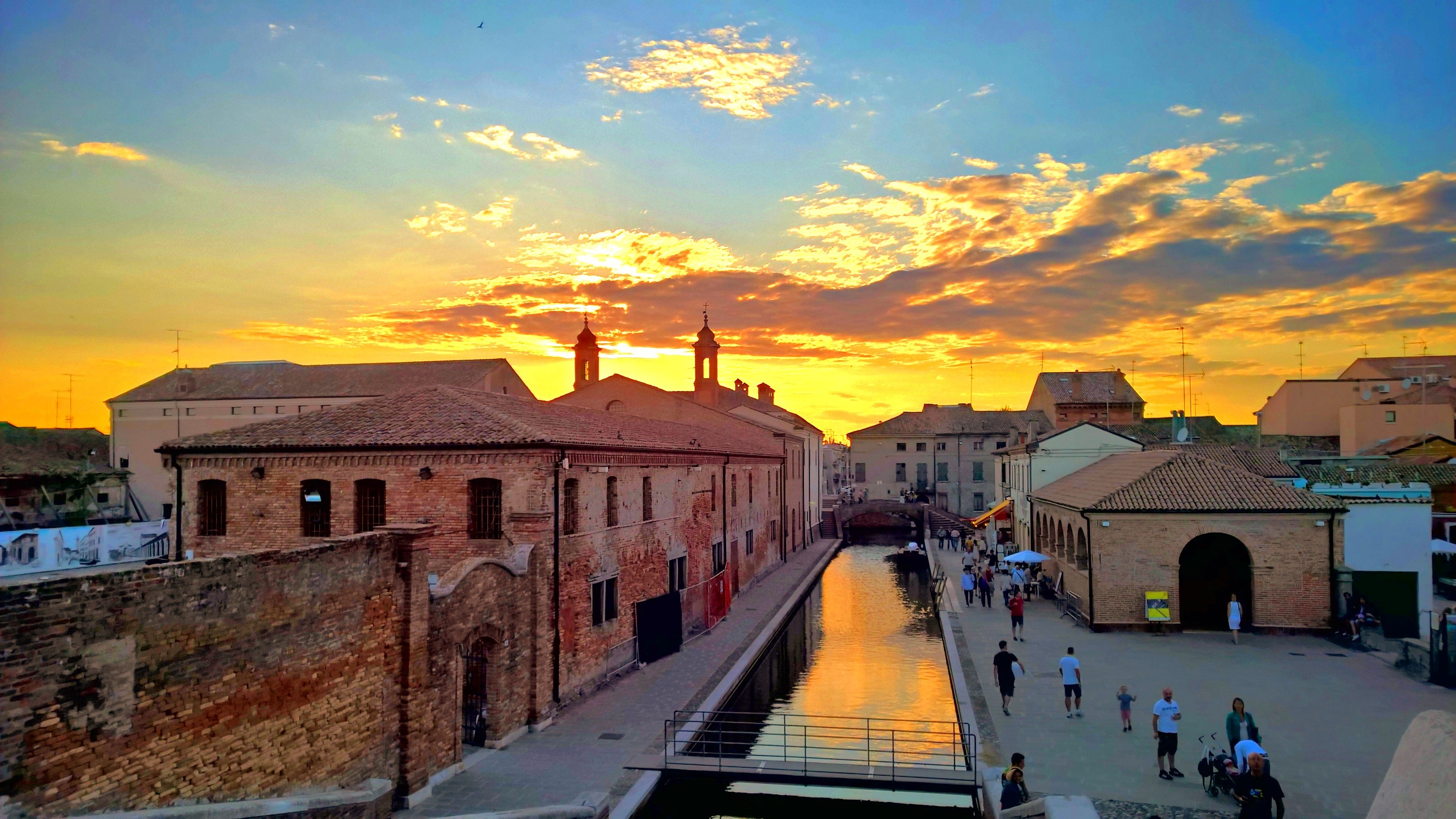 A breathtaking sunset illuminates the sky over Comacchio, Italy, 9 years ago today. Golden clouds stretch across a azure sky, their edges aflame with the sun's dying light. The celestial spectacle is mirrored in the calm waters of a canal running through the heart of the old town. Historic brick buildings line the waterway, their weathered facades glowing warmly in the fading day. Church spires pierce the horizon, silhouetted against the radiant sky. Tourists and locals stroll along the canal's edge, their elongated shadows stretching across the cobblestones, as they savor the magical moment where day gives way to night in this enchanting Italian seaside town.