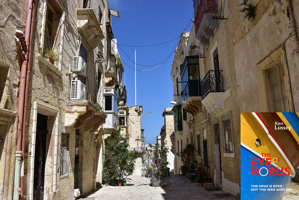 This modern image shows a view along a street in Senglea, Malta. Three-storey houses built of a light honey-coloured stone line both sides of the street, which is paved. There are balconies protruding from many upper floor rooms and the right side of the street is in shadow. The street continues into the frame past a mass of plants in pots and then descends steeply, out of sight. The sky is blue. The front cover of ‘The Eye of Horus’ is shown in the bottom right corner.